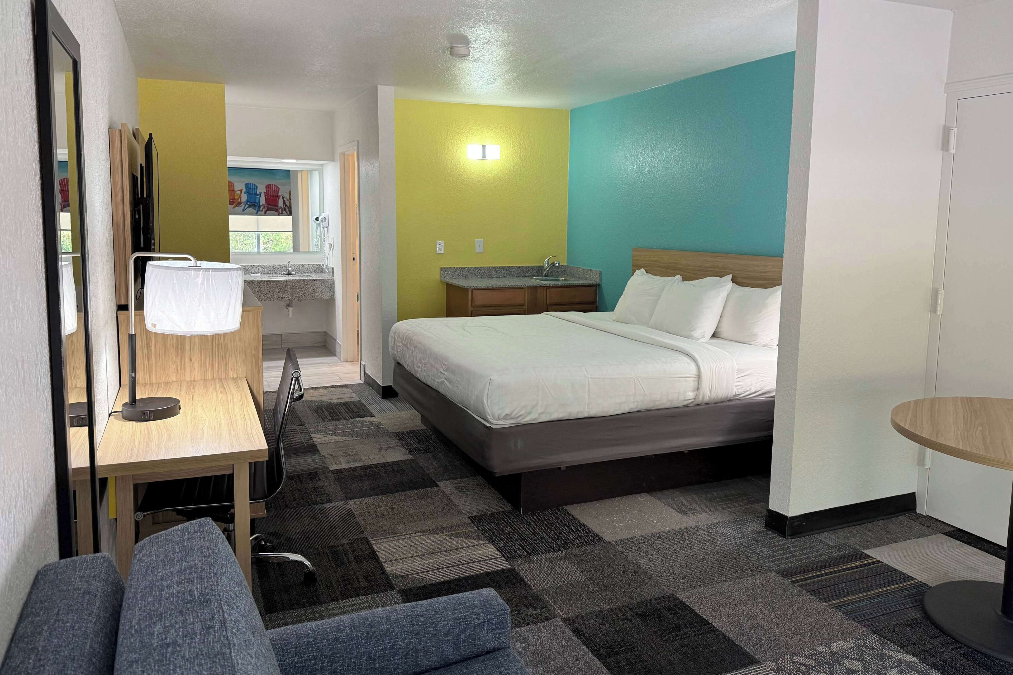 A hotel room with a bed, a desk with a lamp, a small bathroom with a sink, and a view of the outside.