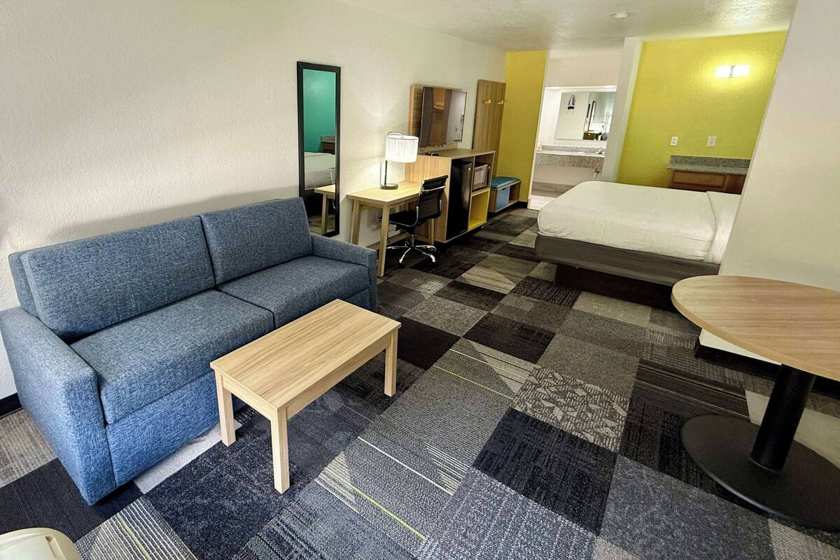 A hotel room with a blue sofa, a wooden coffee table, a desk with a computer, a bed, and a small table.