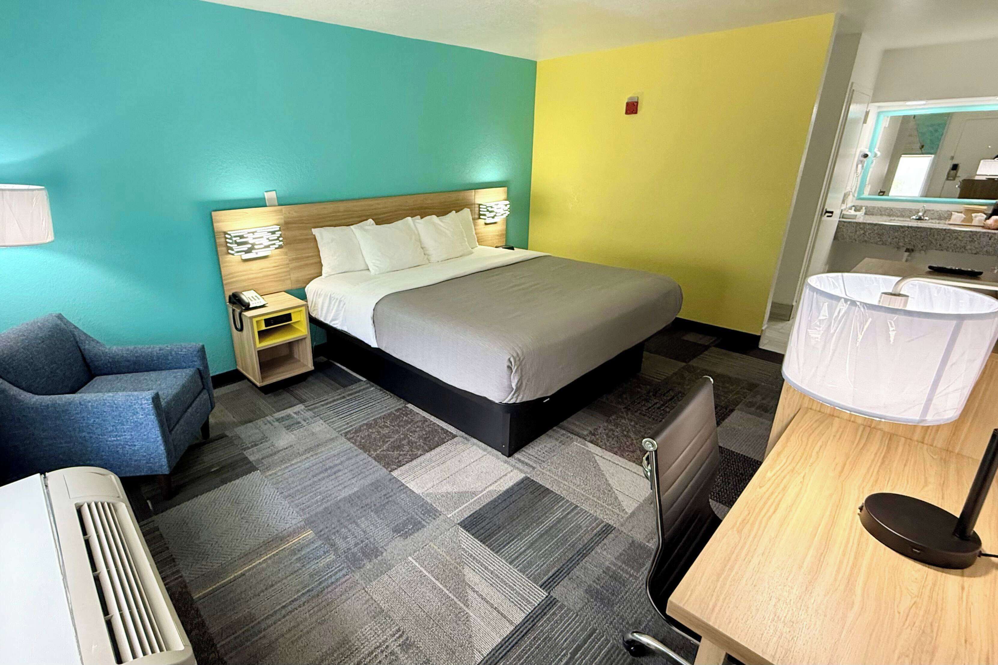 A hotel room with a bed, bedside table, chair, desk, and a bathroom with a sink and mirror.