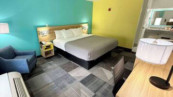 A hotel room with a bed, bedside table, chair, desk, and a bathroom with a sink and mirror.
