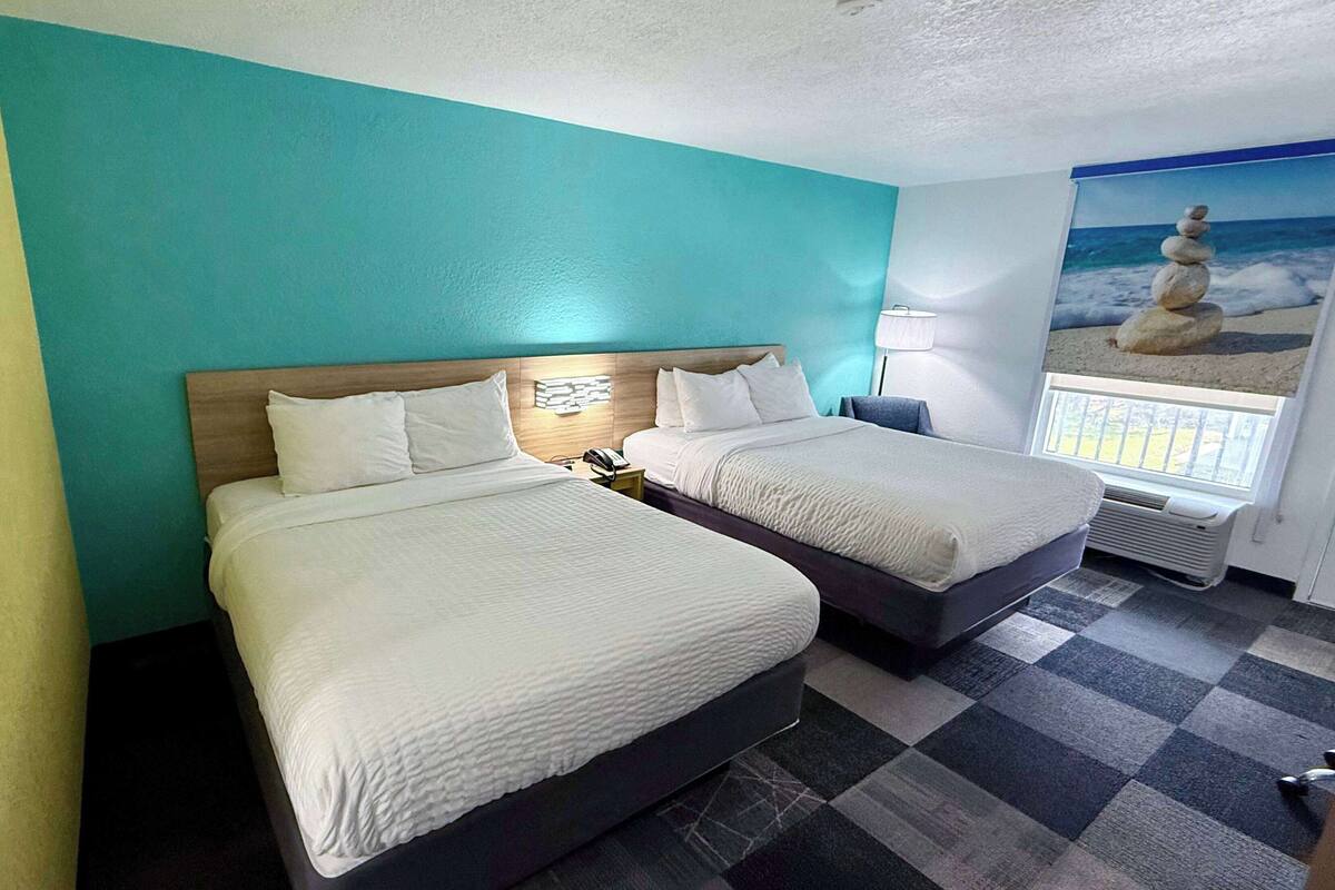 A hotel room with two beds, a check-in counter, a window with a view of the beach, and a wall-mounted air conditioning unit.