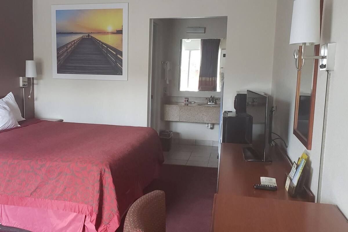 A hotel room with a bed, a desk, a TV, a bathroom visible through an open door, and a sunset painting on the wall.