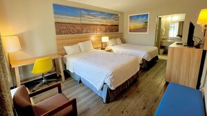 Une chambre d’hôtel avec deux lits, un bureau avec une chaise, une petite table, un miroir et des œuvres d’art accrochées aux murs.
