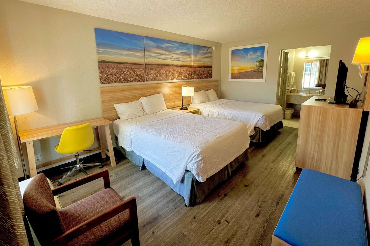A hotel room with two beds, a desk with a chair, a small table, a mirror, and artwork on the walls.