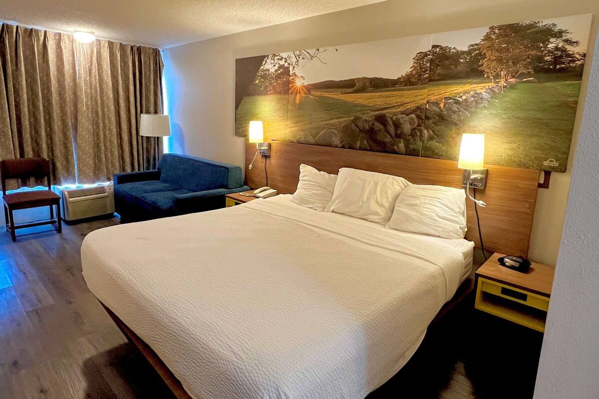 A hotel room with a large bed, a blue armchair, a wooden nightstand with a lamp, and a painting of a landscape on the wall.