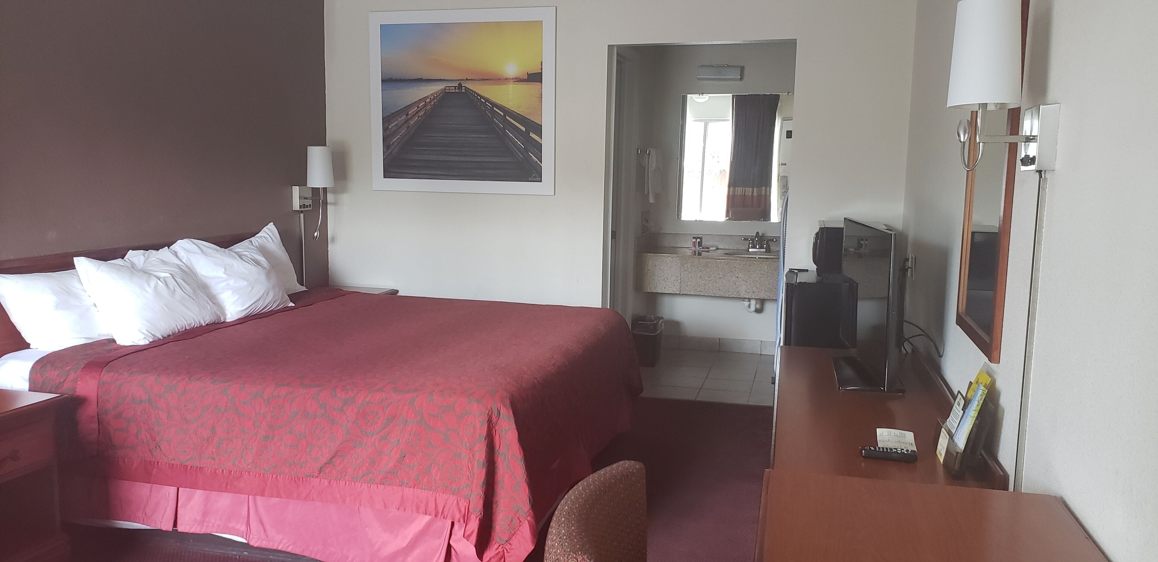 A hotel room with a large bed, a desk with a chair, a television, a bathroom with a sink and mirror, and a painting of a pier on the wall.