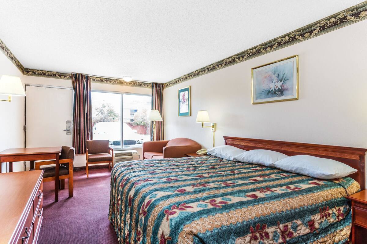 A hotel room with a bed, a desk, a chair, a window with curtains, and a painting on the wall.