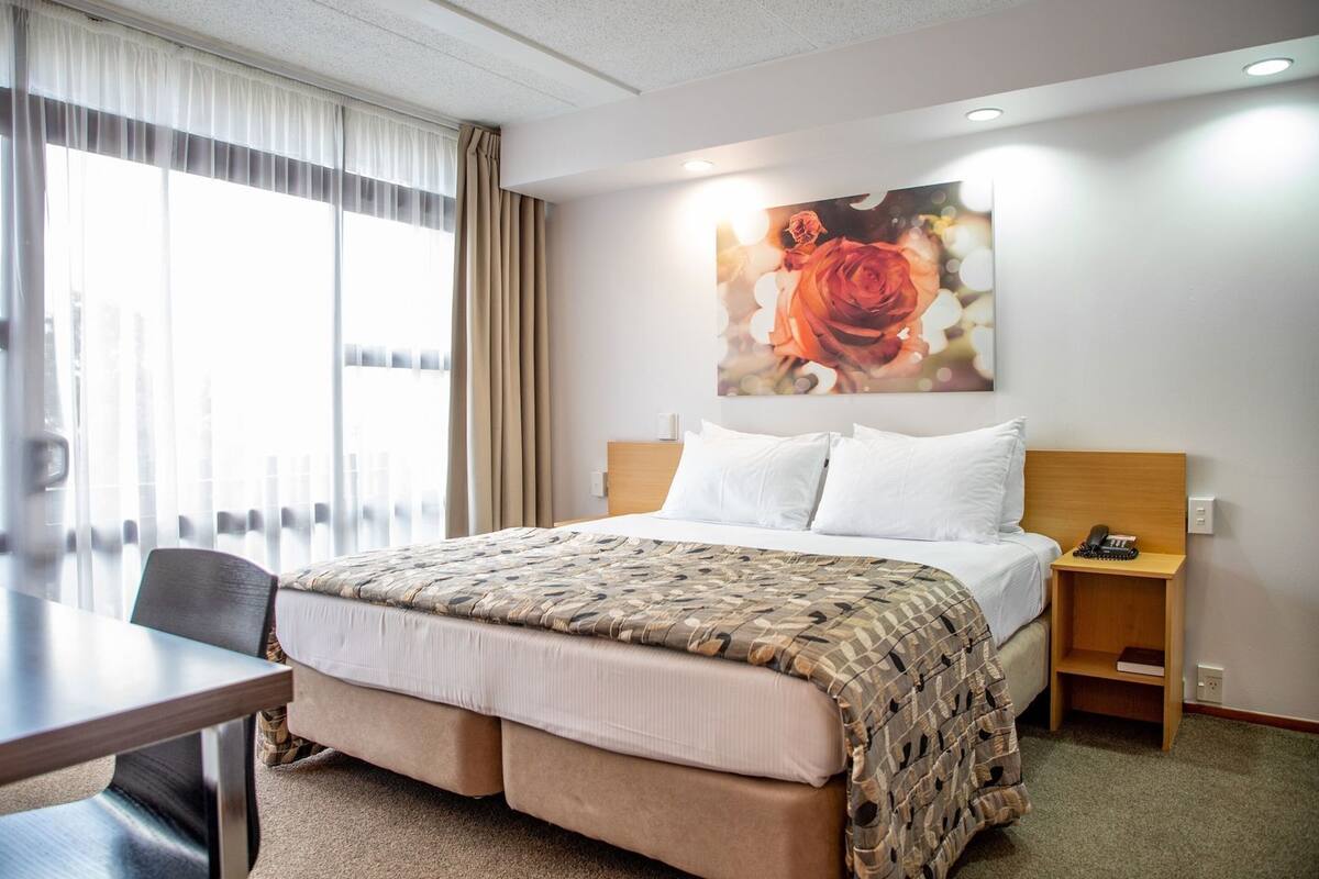A hotel room with a bed, a desk, a chair, a nightstand, and a wall-mounted painting.