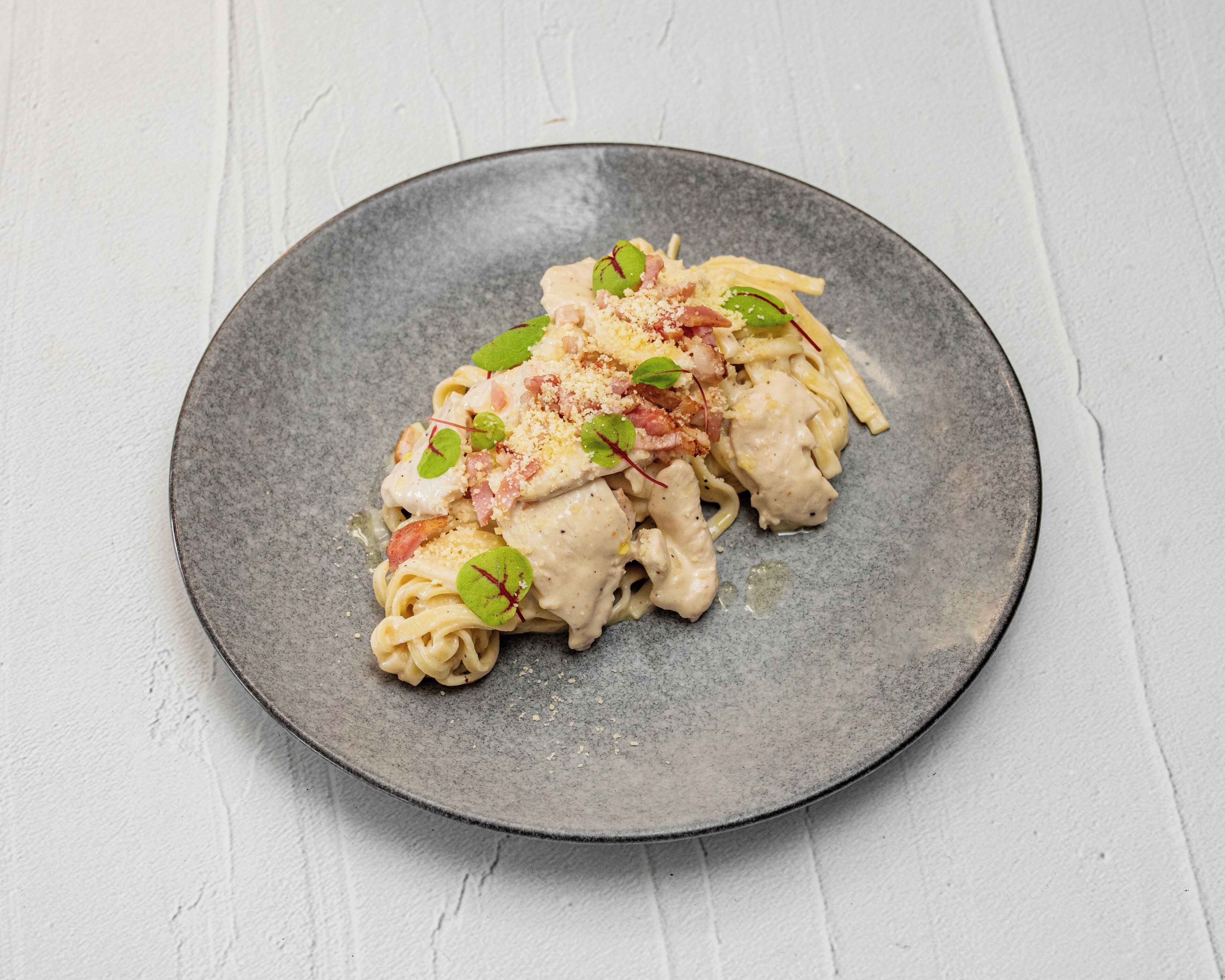 Une assiette de pâtes nappées d’une sauce crémeuse, garnie de feuilles vertes et de morceaux de lardons.