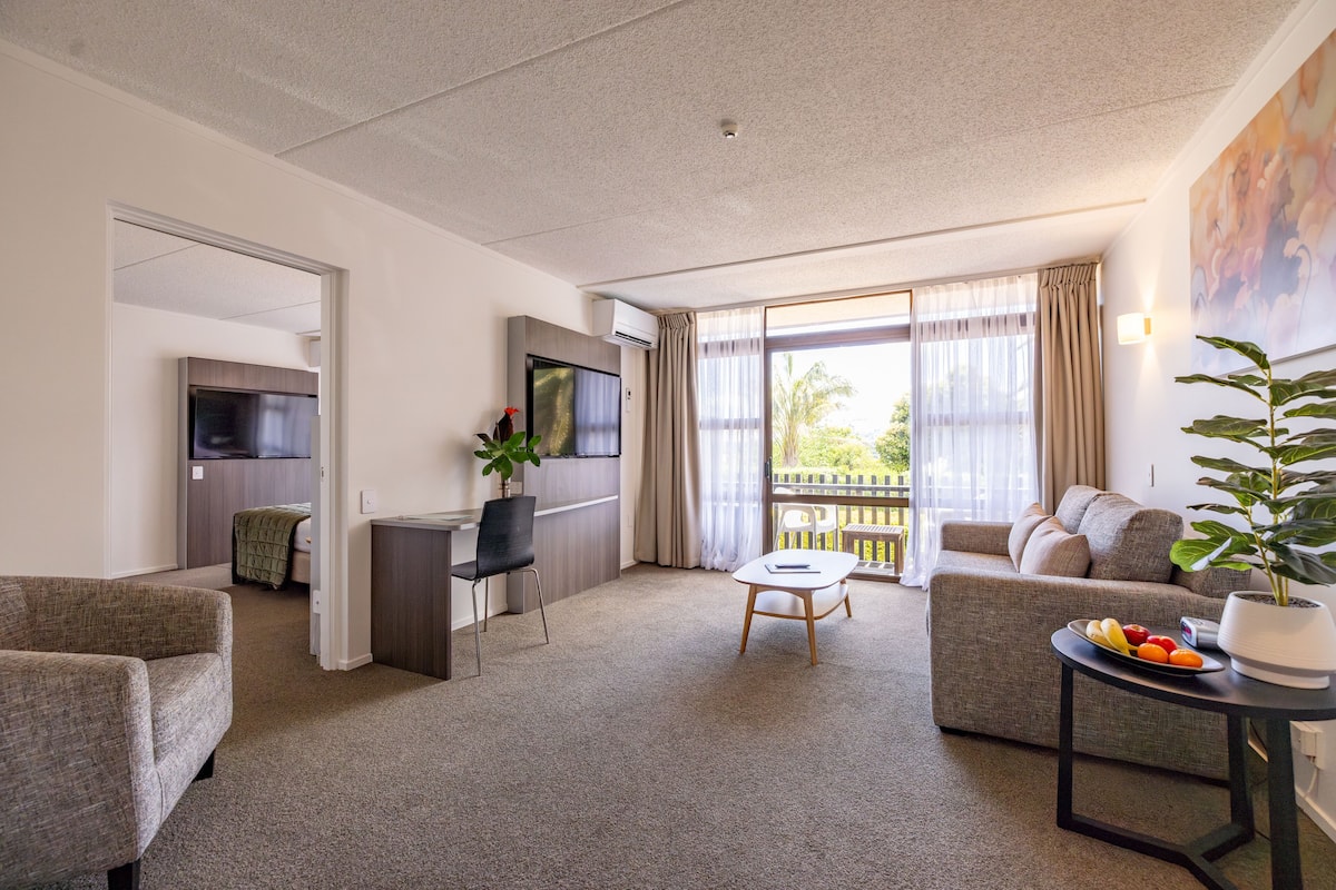 A modern living room with a sofa, a dining table, and a TV. There is a balcony with a view of palm trees.
