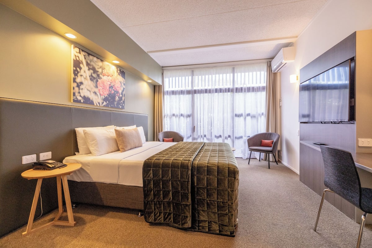 A hotel room with a bed, a desk, a chair, a TV, and a window with curtains.