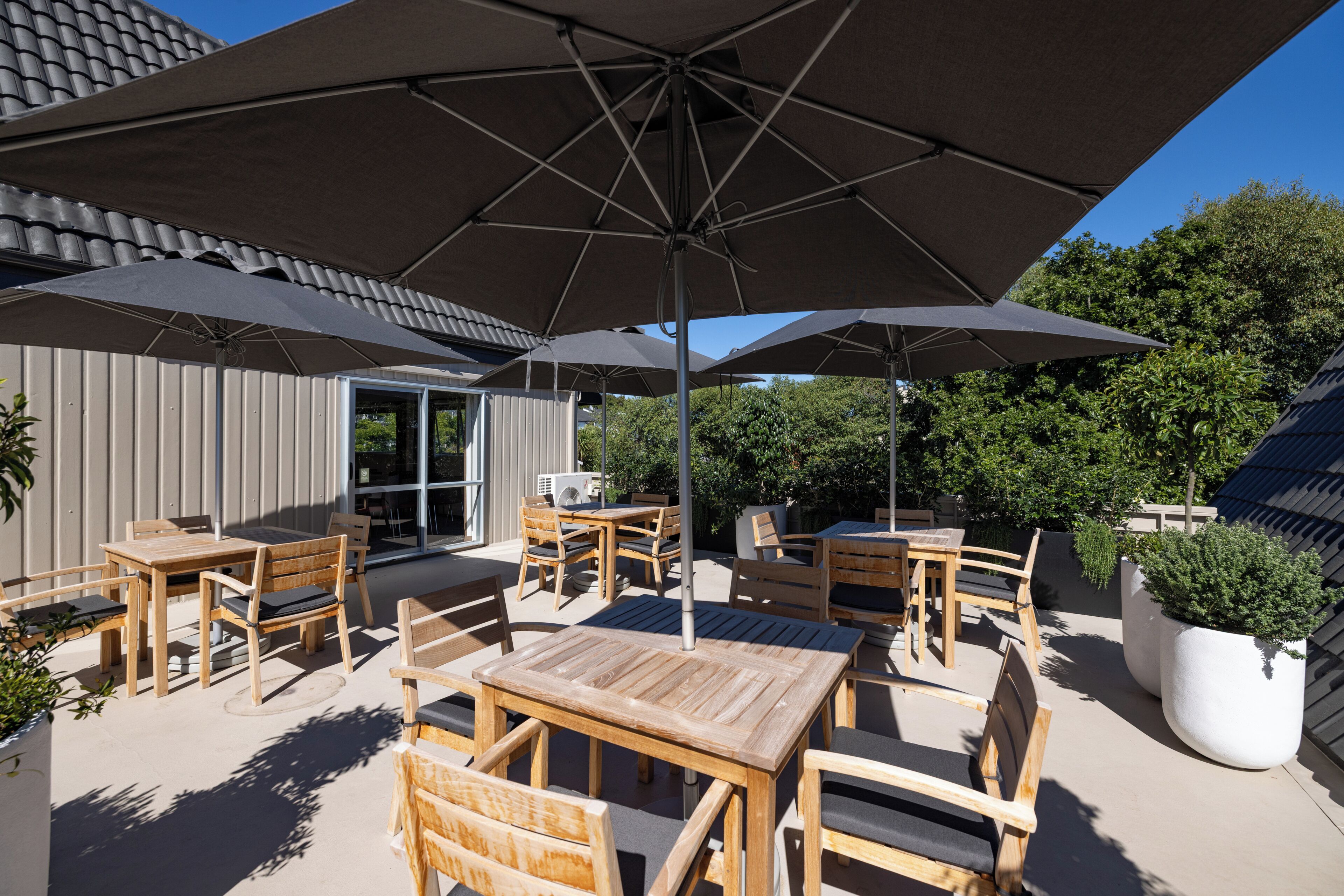 An outdoor patio area with wooden tables and chairs, large umbrellas, and a potted plant.
