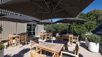 An outdoor patio area with wooden tables and chairs, large umbrellas, and a potted plant.