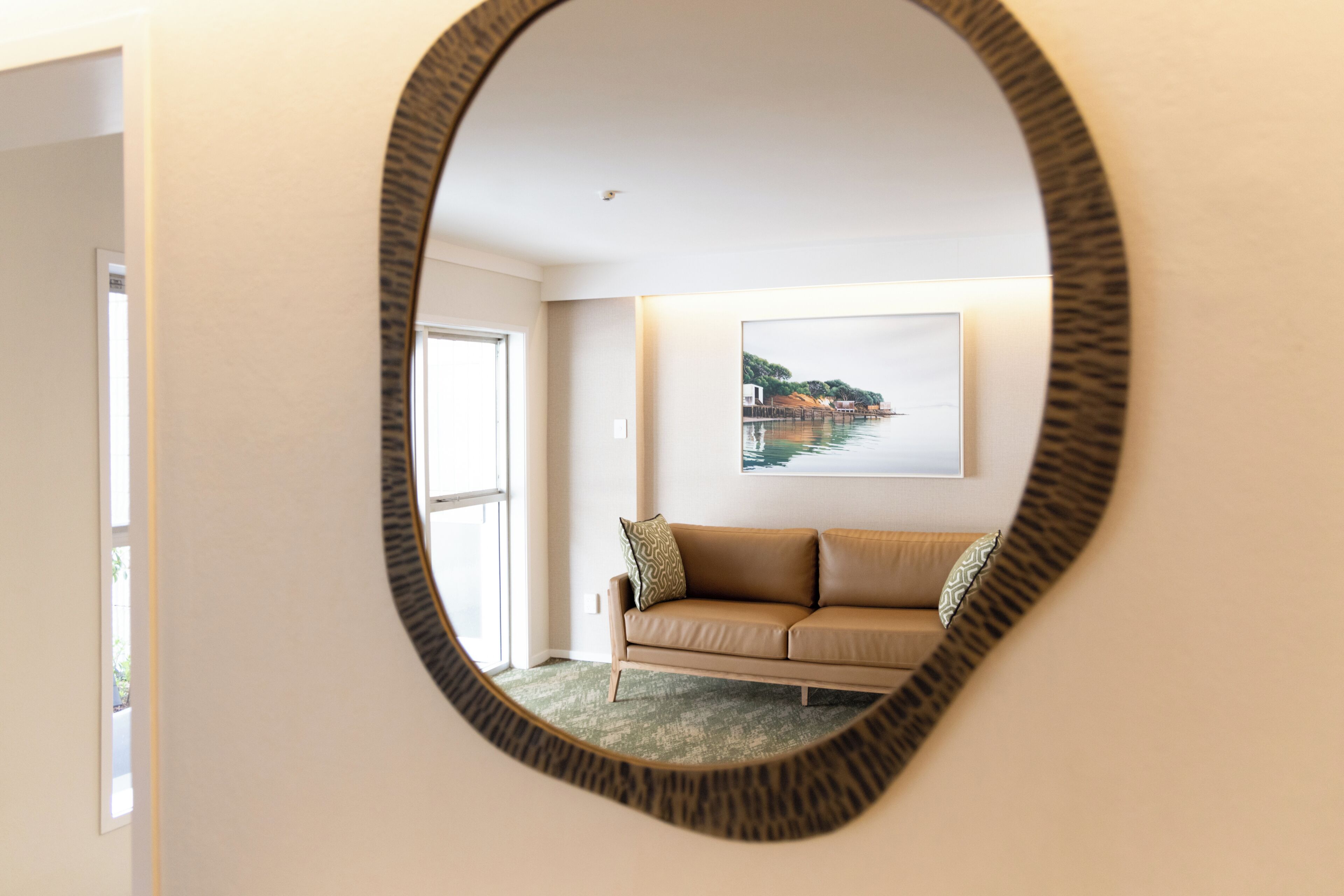 A room with a circular mirror reflecting a beige sofa and a framed picture of a coastal landscape.
