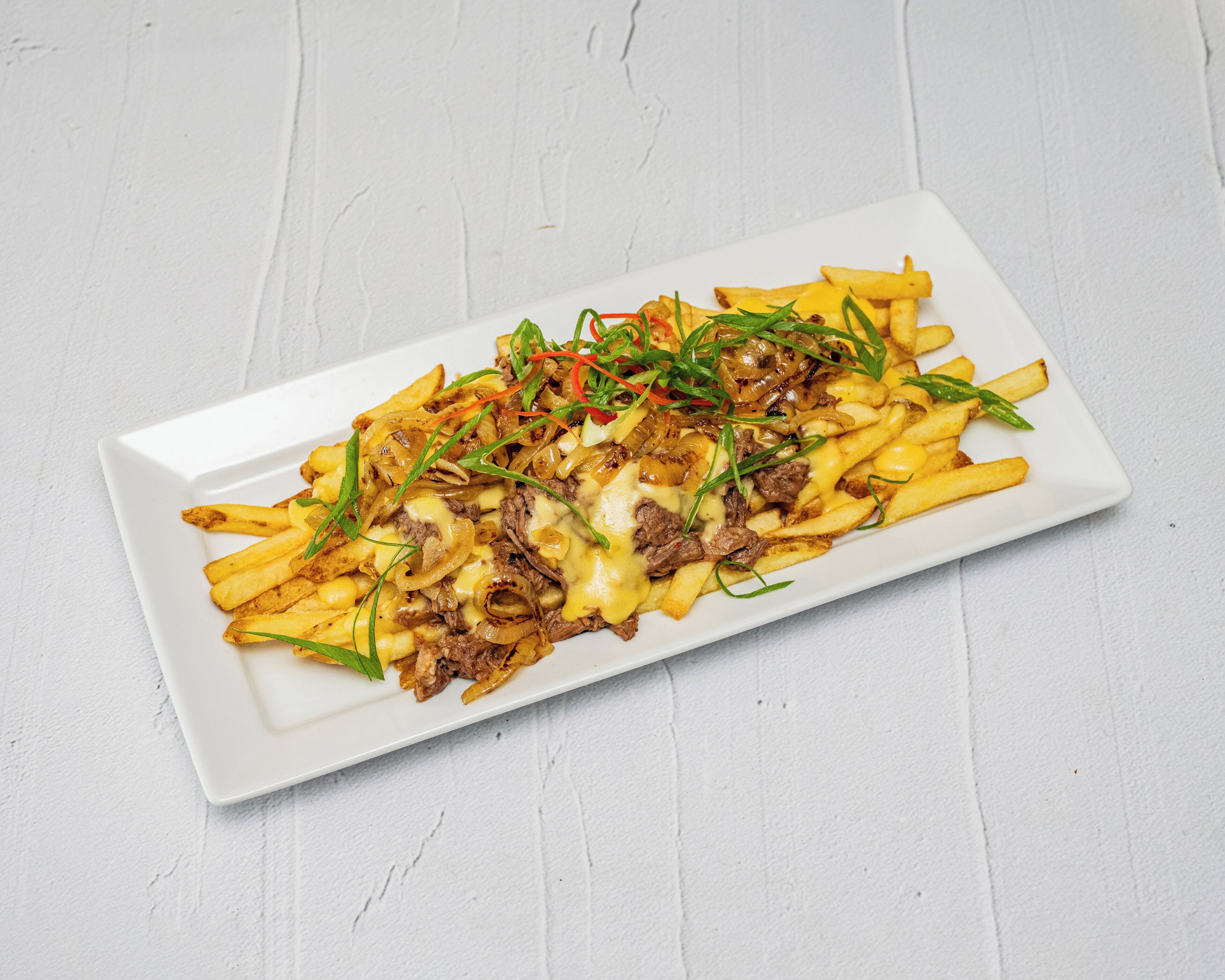 Une assiette rectangulaire blanche contenant une portion de frites recouverte de fromage fondu, d’oignons et d’herbes vertes.