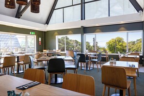 A spacious dining area with multiple tables and chairs, large windows offering a view of the outdoors, and a high ceiling with hanging lights. - Auckland Rose Park Hotel (Auckland)