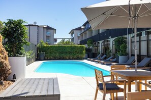 Una piscina con sillas de descanso y sombrillas, rodeada por un seto y un edificio con balcones.