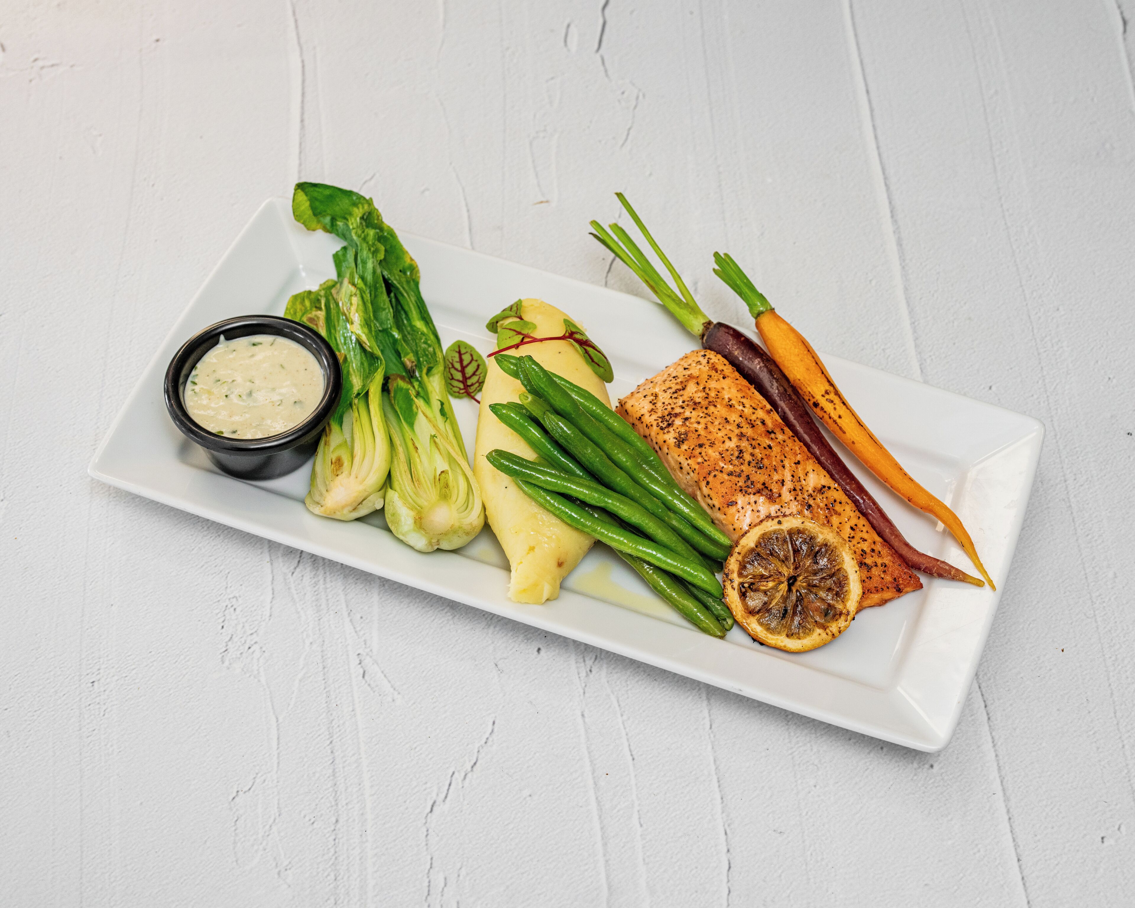 Une assiette rectangulaire blanche contenant divers aliments, notamment un morceau de saumon, des haricots verts, des carottes et une portion de purée de pommes de terre.