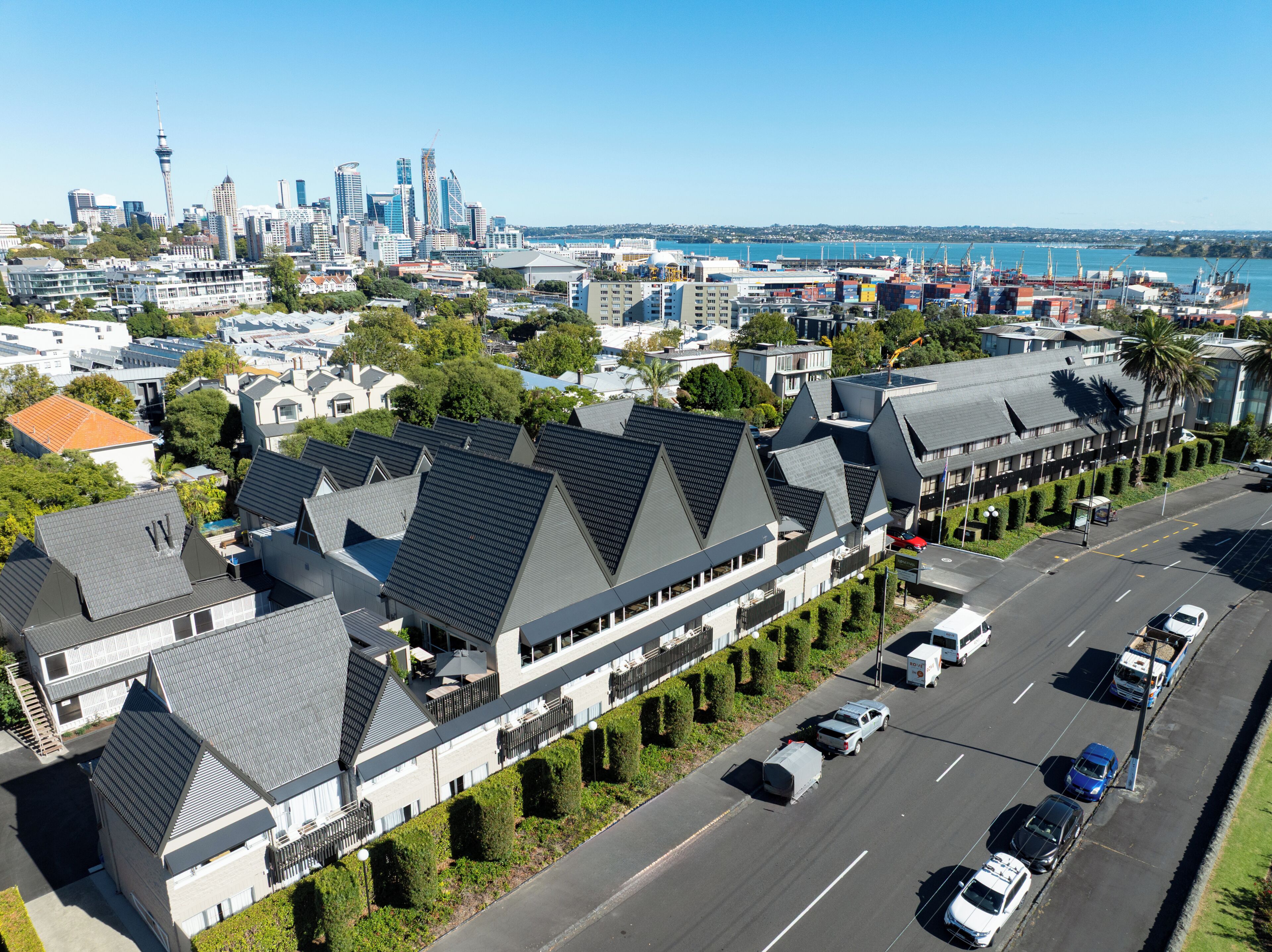Un hôtel moderne, avec une série de chambres caractérisées par des toits sombres et des murs blancs, situé dans une ville offrant une vue sur les grat-ciel et un port de plaisance.