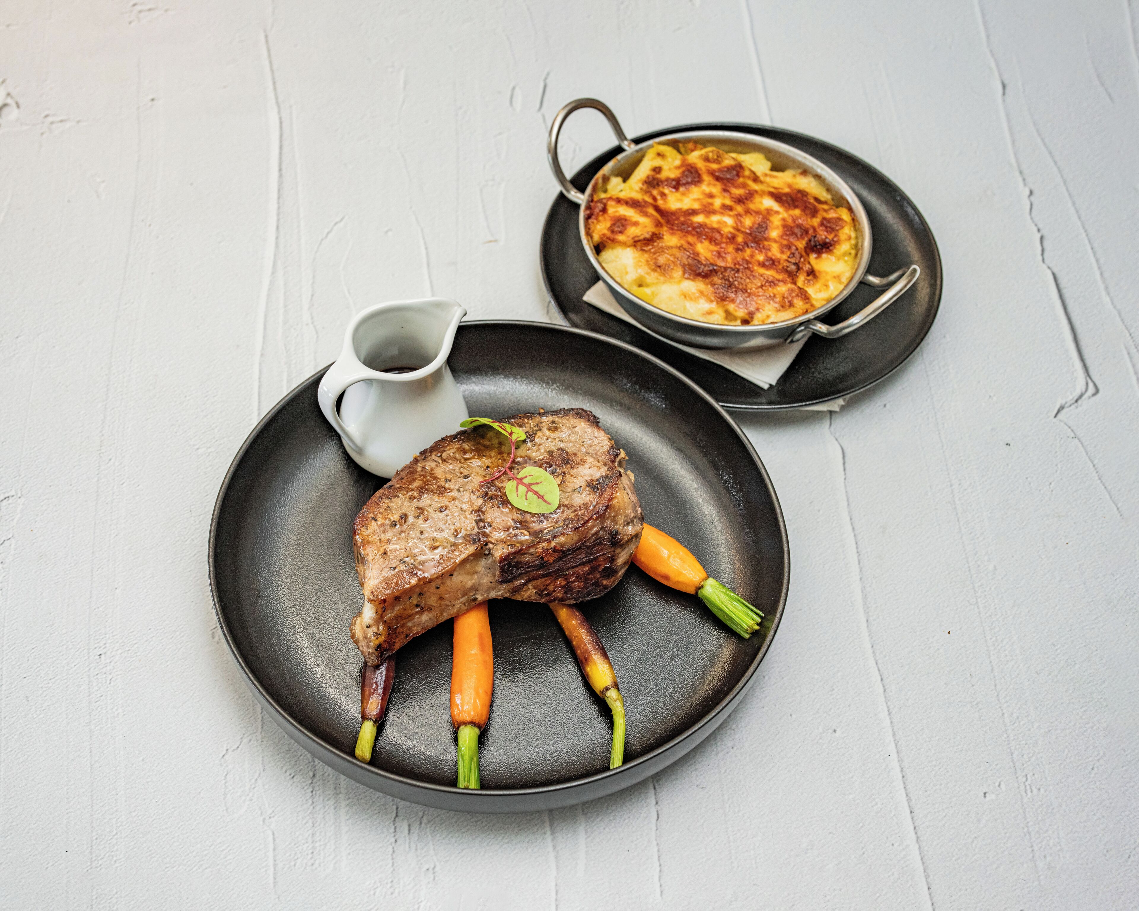 A steak with carrots on a plate, a bowl of baked potatoes, and a small pitcher of sauce.