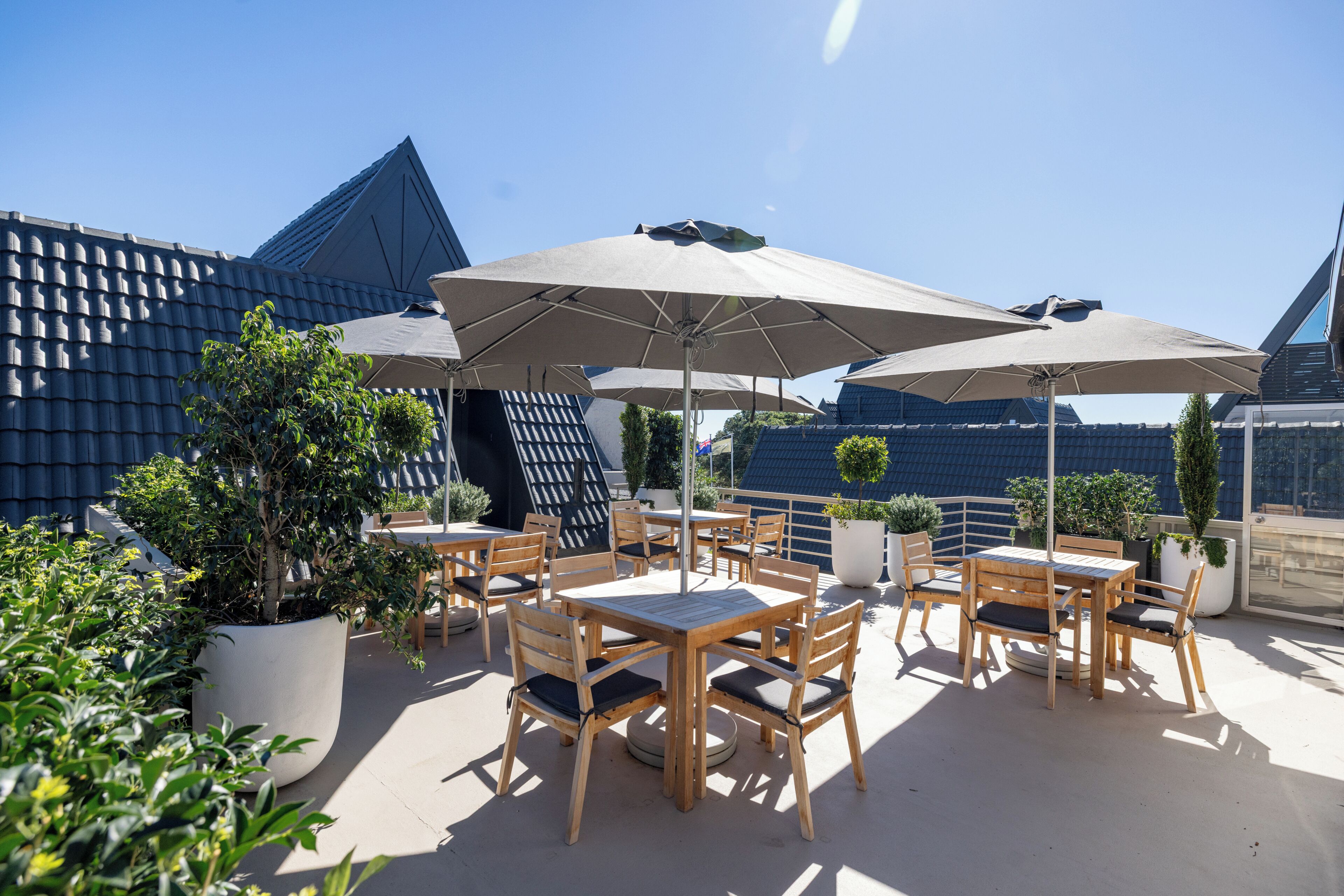 Eine Terrasse im Freien mit Holztischen und -stühlen, großen Sonnenschirmen und Blumentöpfen.