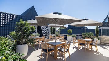 Eine Terrasse im Freien mit Holztischen und -stühlen, großen Sonnenschirmen und Blumentöpfen.