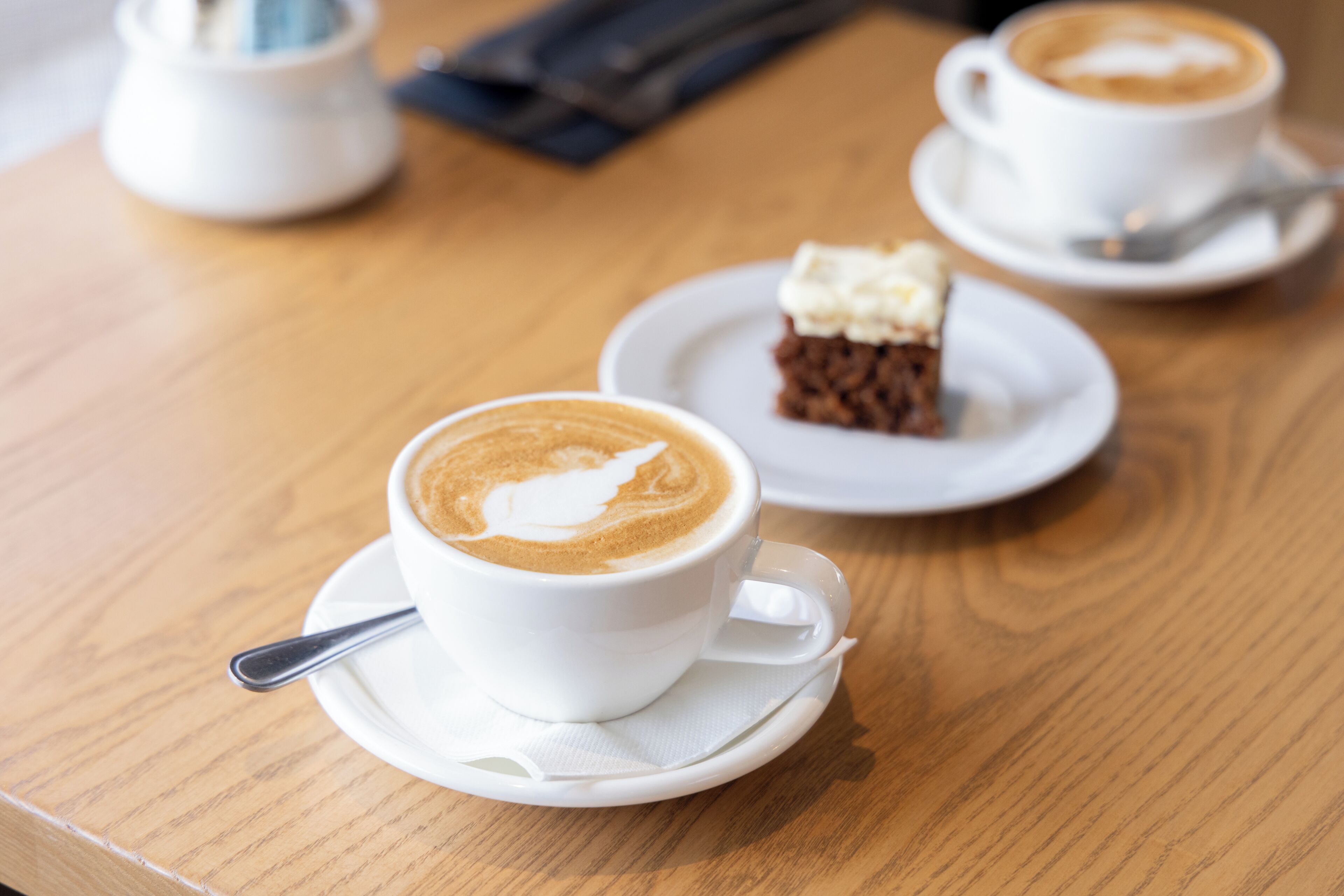 A wooden table with two white cups of coffee and a slice of cake on a saucer.
