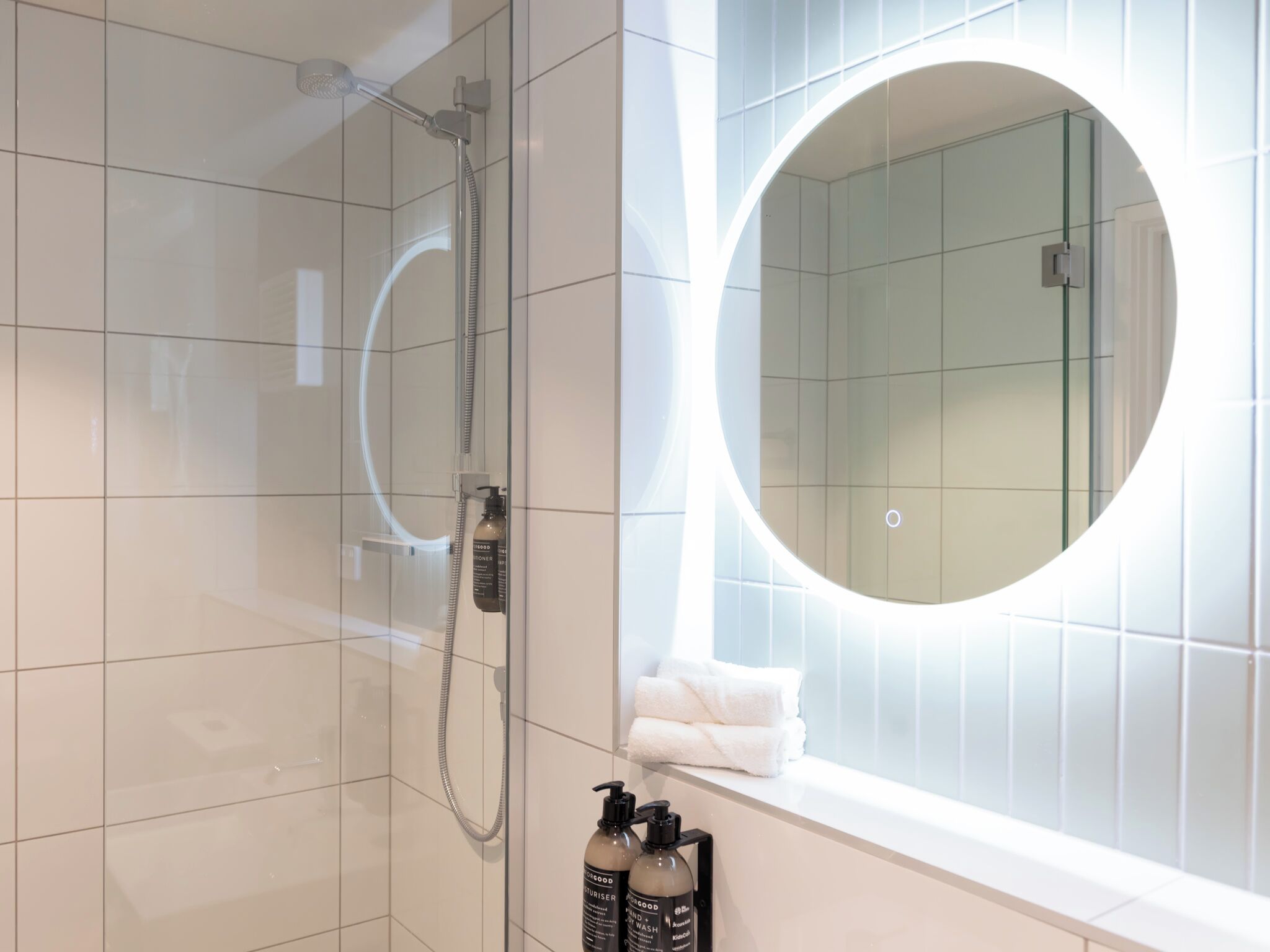 A modern bathroom with a glass-enclosed shower, a round mirror, and toiletries on the wall.