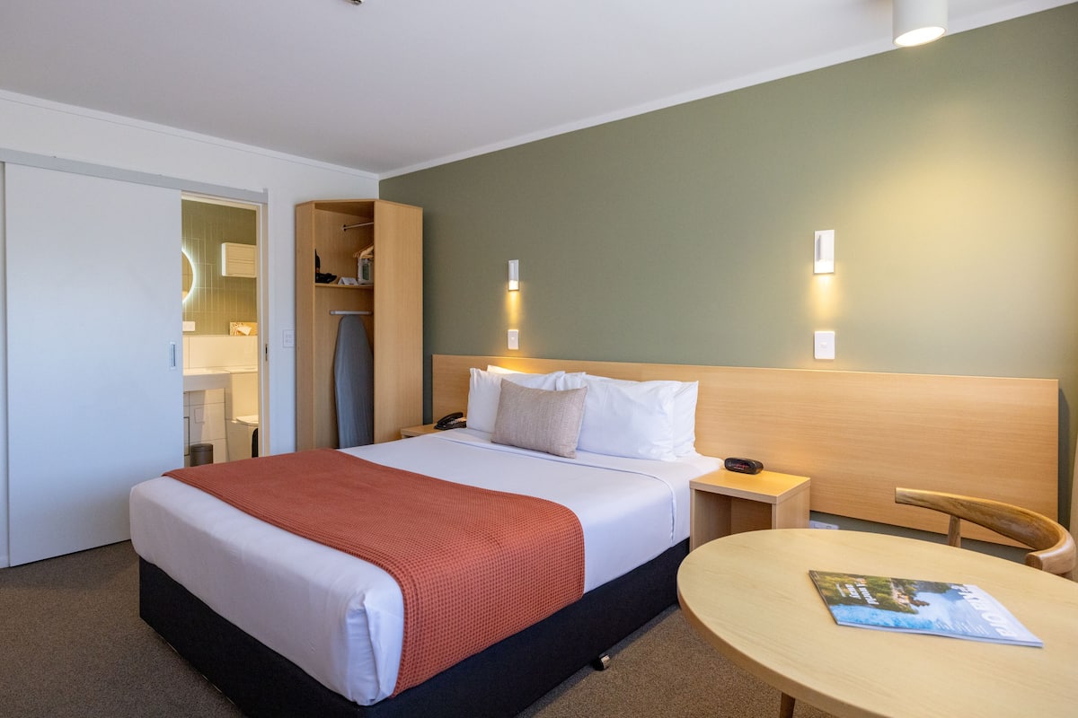 A hotel room with a bed, bedside table, chair, and a round table with a magazine.