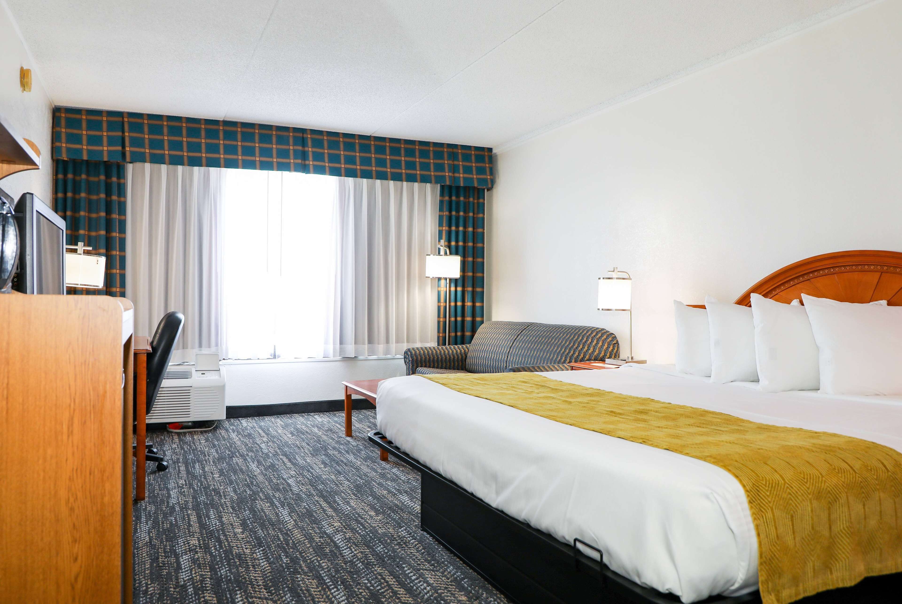 Une chambre d’hôtel avec un lit, un bureau, une chaise, une lampe et une fenêtre avec des rideaux.