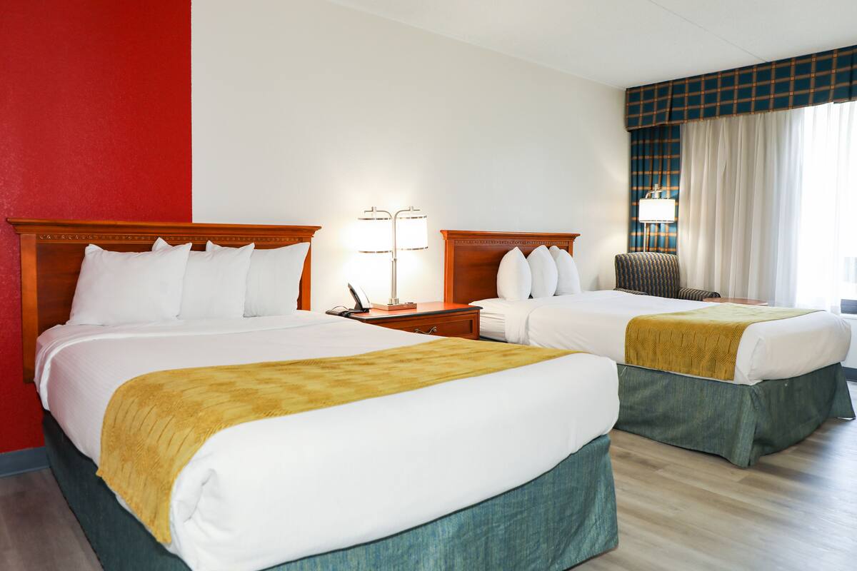 Habitación de hotel con dos camas, una mesita de noche con lámpara, una silla y un ventanal con cortinas.