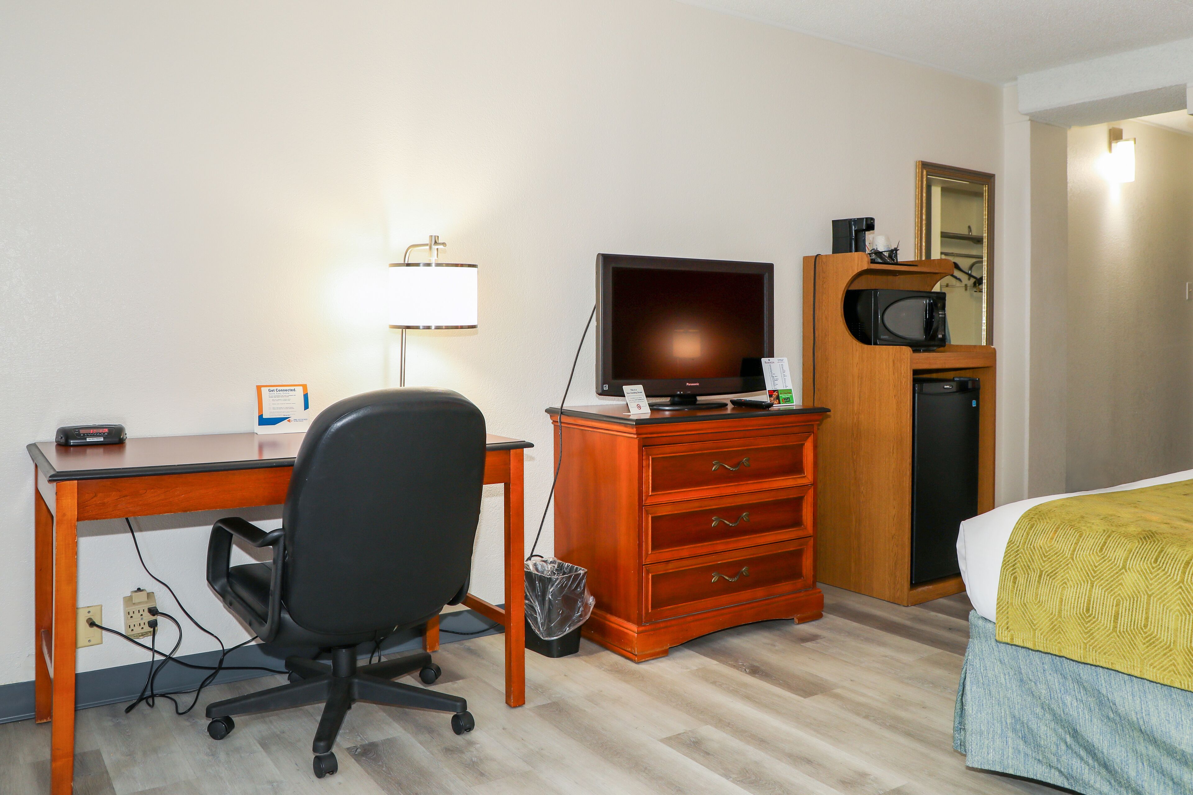 Habitación de hotel con escritorio de madera, silla de oficina negra, televisor sobre una cómoda de madera, microondas y una cama con manta amarilla.
