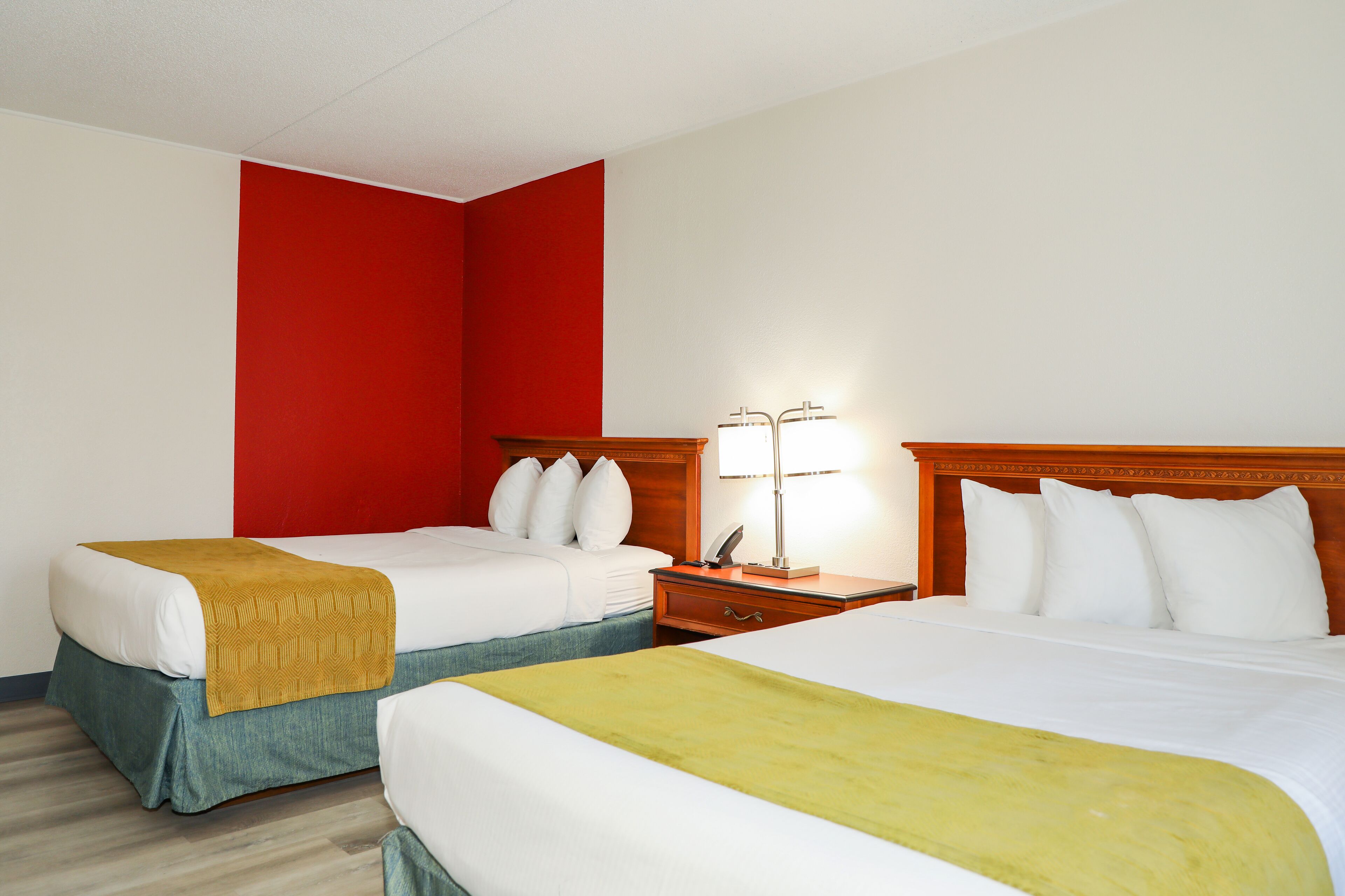 Habitación de hotel con dos camas, un cabecero de madera, una mesita de noche con lámpara y una pared de acento en rojo.
