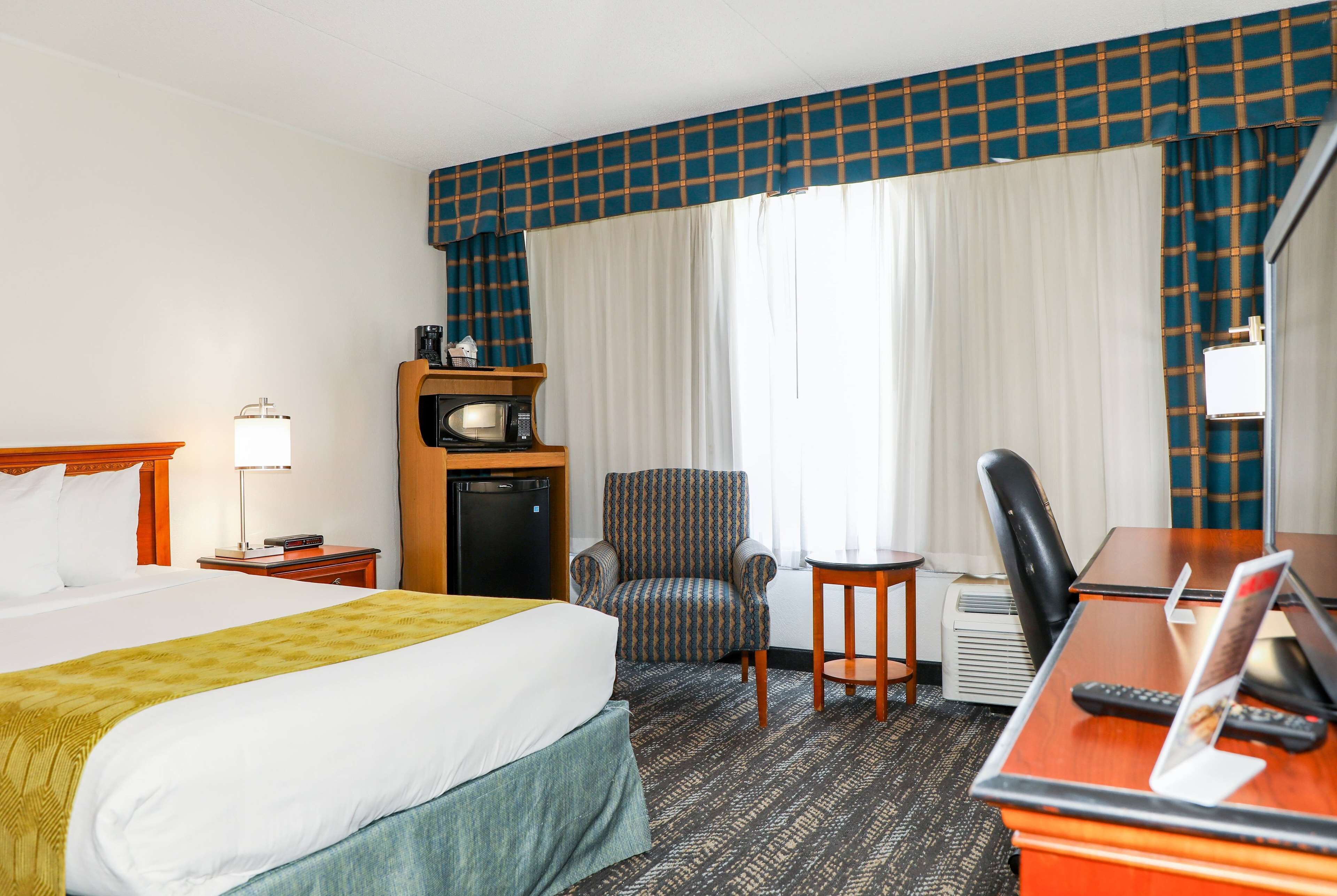 Quarto de hotel com cama, escrivaninha com computador, cadeira, mesinha, micro-ondas e televisão.