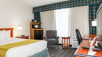 Quarto de hotel com cama, escrivaninha com computador, cadeira, mesinha, micro-ondas e televisão.