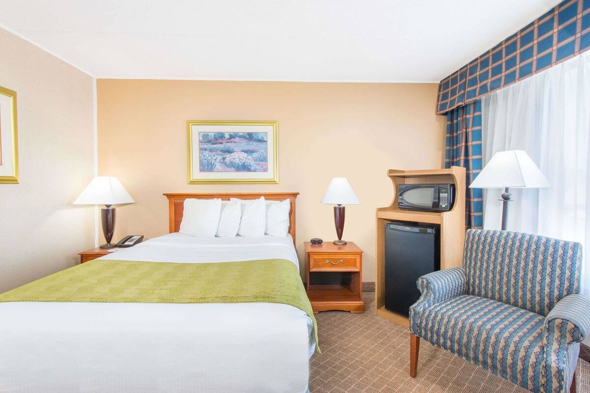 Habitación de hotel con una cama, dos lámparas de noche, una silla, una mesita con lámpara, un microondas y un cuadro en la pared.