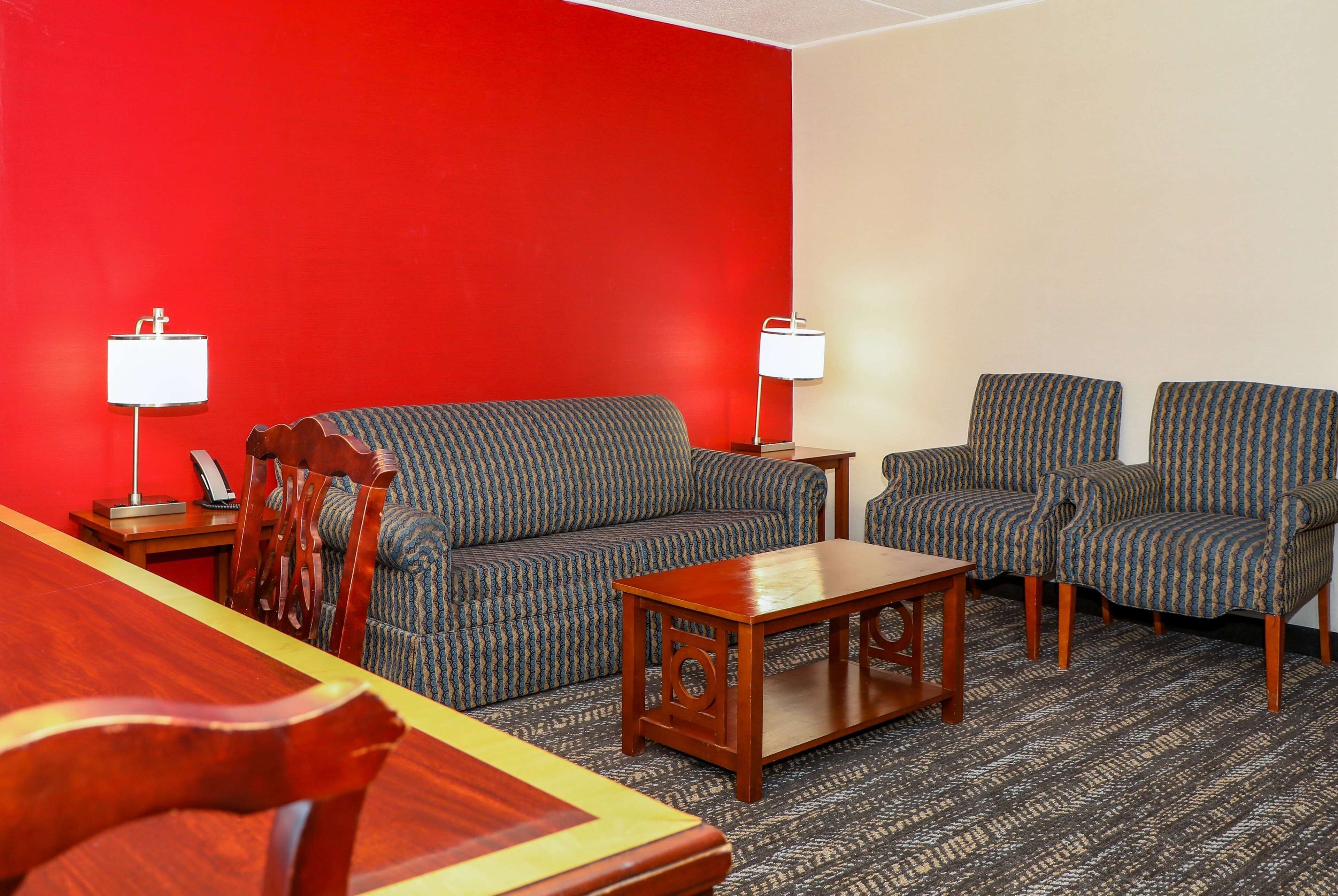 A hotel room with a red accent wall, a patterned sofa, two armchairs, a wooden coffee table, and two bedside lamps.