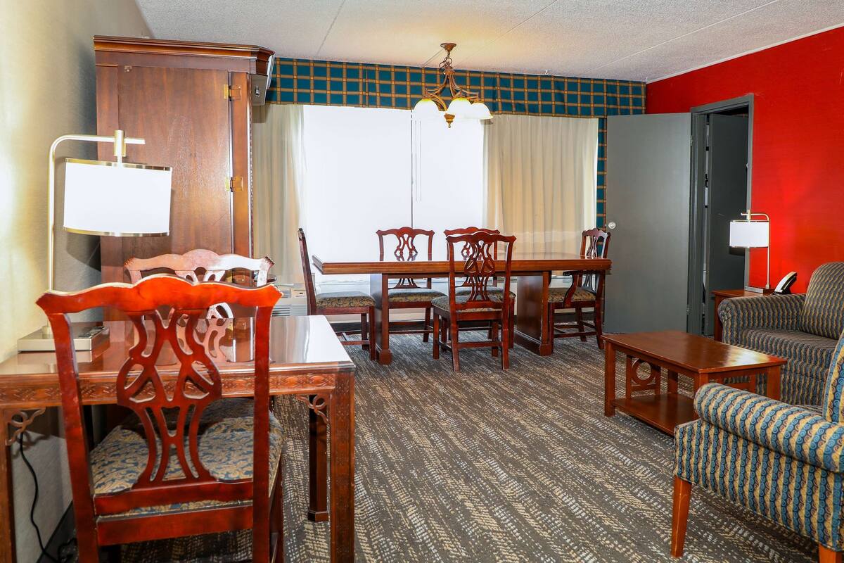 Habitación de hotel con una mesa de comedor, cuatro sillas, un sofá, una lámpara y un ventanal con cortinas.