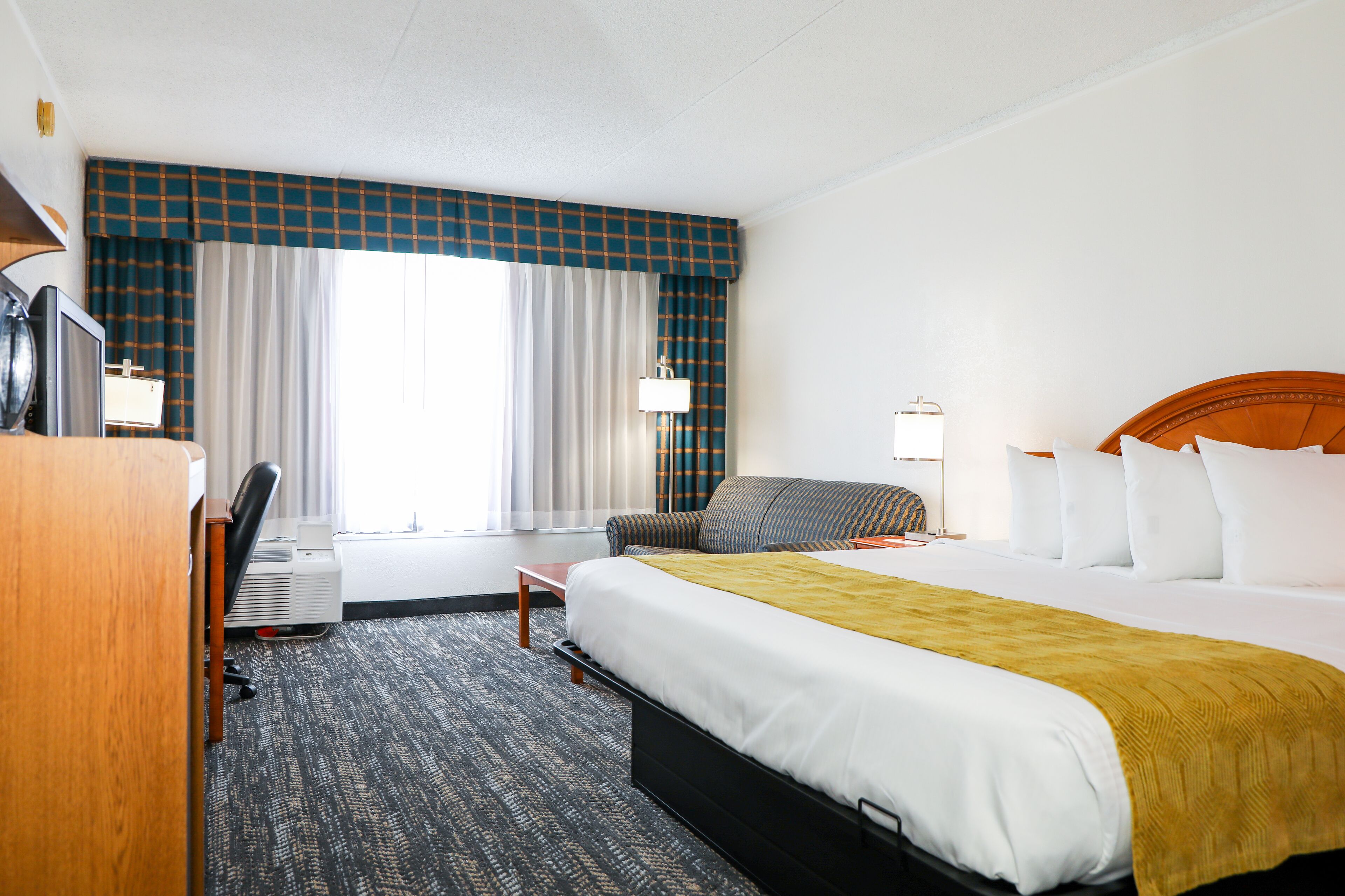 A hotel room with a bed, a desk, a chair, a lamp, and a window with curtains.