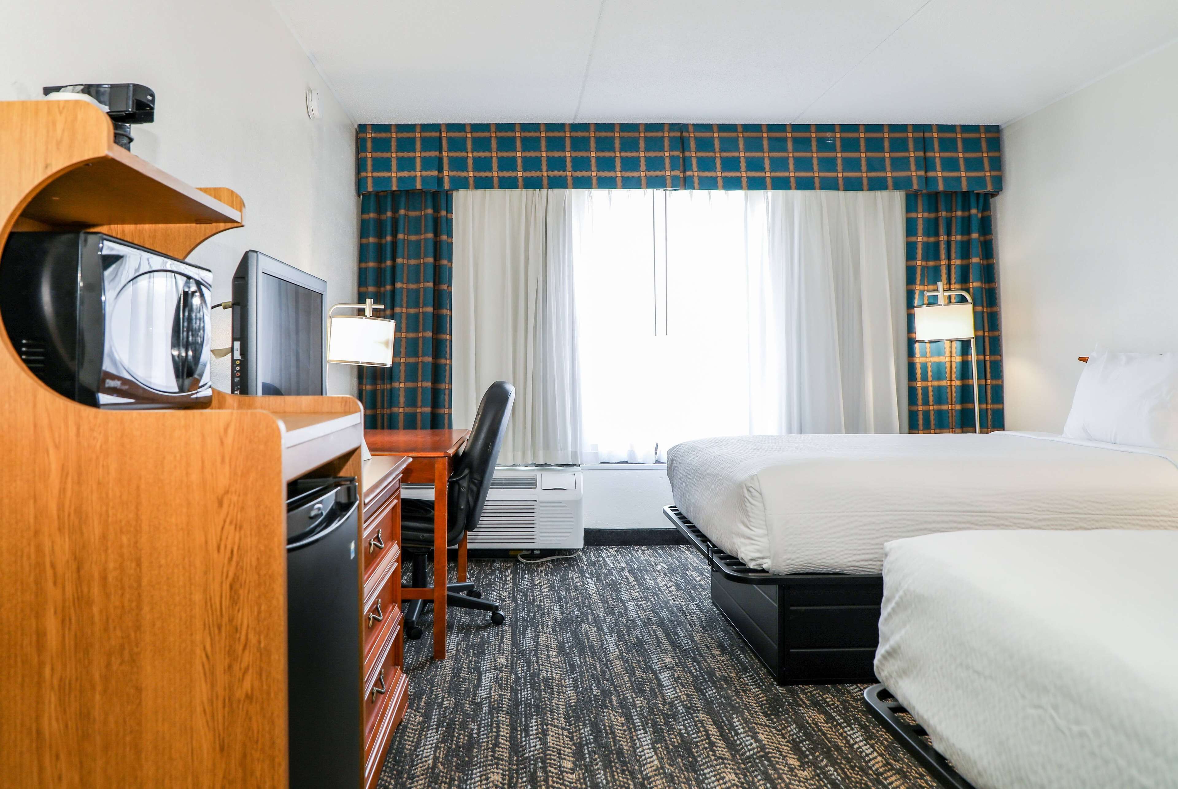 Quarto de hotel com uma cama, uma secretária com televisão, uma cadeira e uma janela com cortinas.