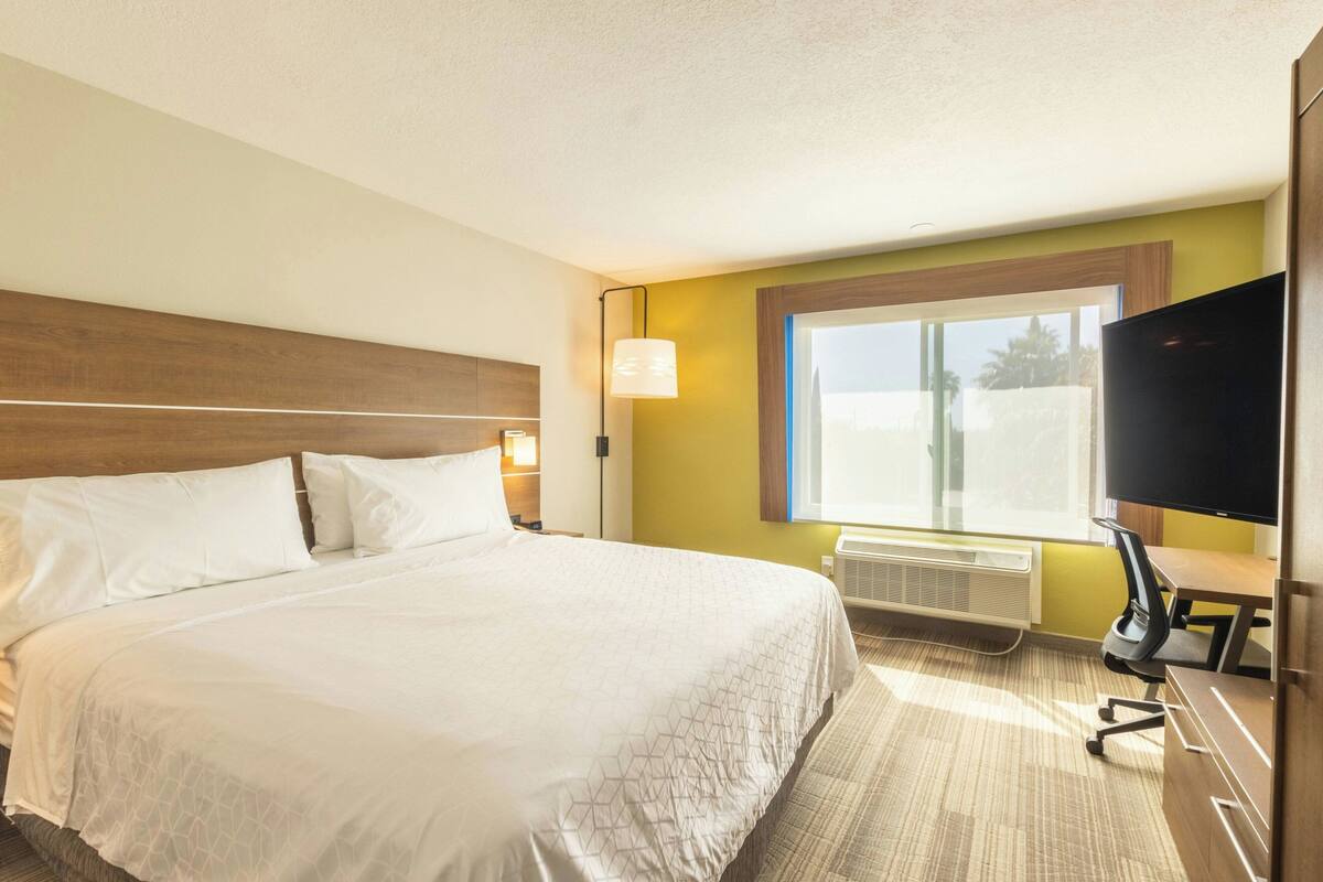 A hotel room with a large bed, a desk with a chair, a television, and a window with blinds.