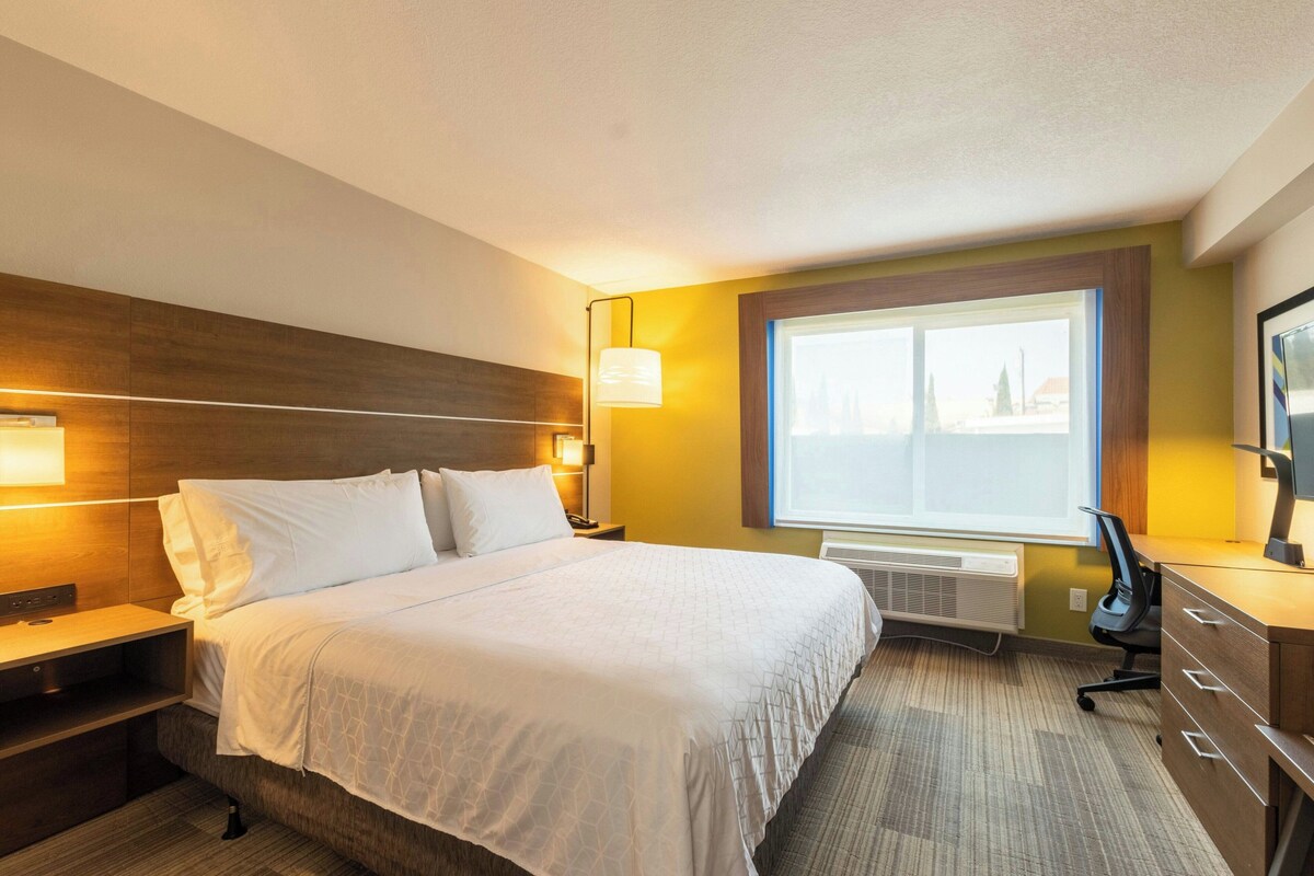 A hotel room with a bed, bedside tables, a desk, a chair, a window, and a wall-mounted lamp.