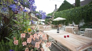 Un patio al aire libre con mesas y sillas de madera, rodeado de exuberante follaje y plantas con flores.