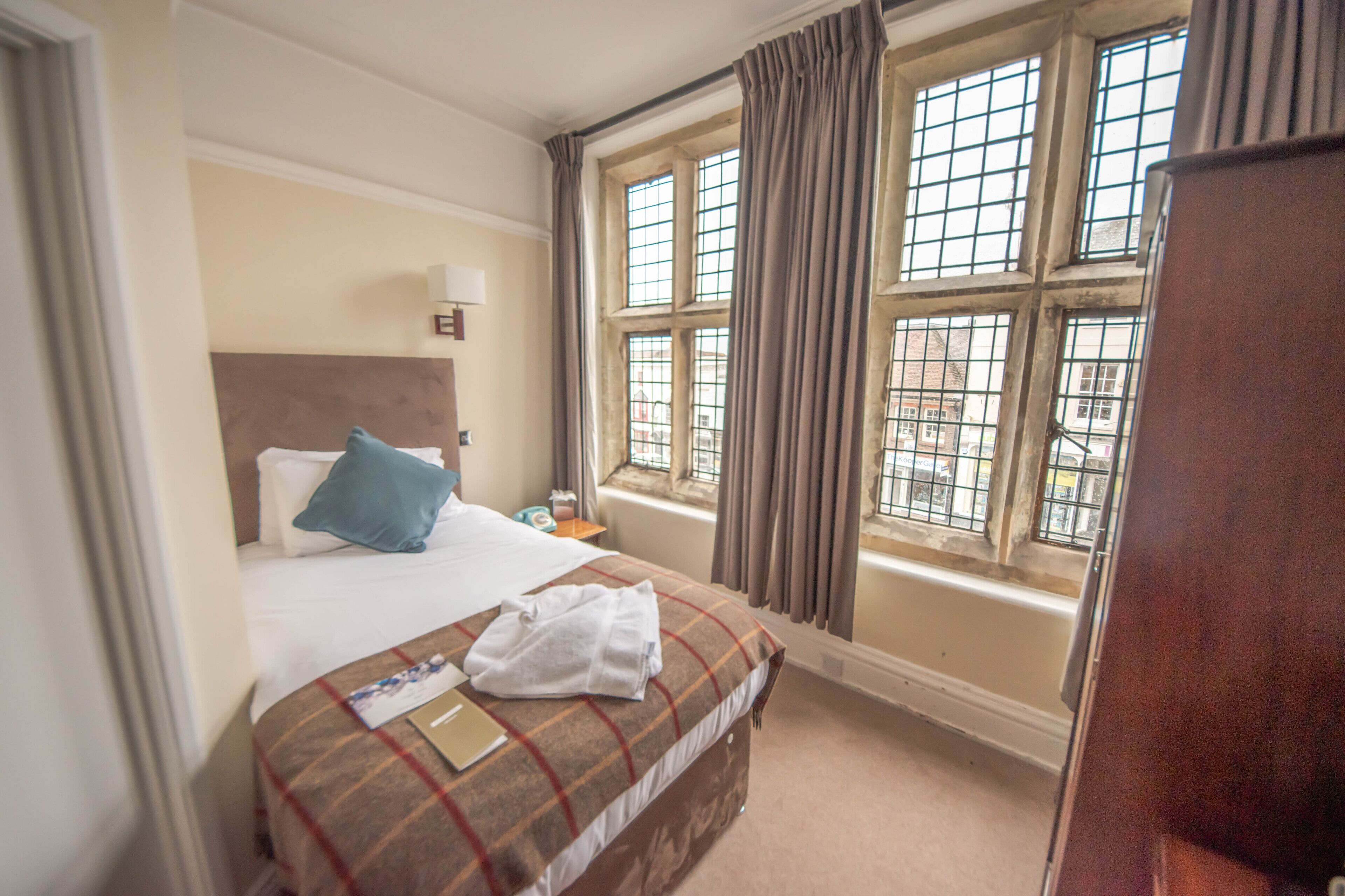 A hotel room with a bed, a window with curtains, a bedside table, and a lamp.