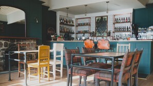 Zona de bar con mesas y sillas de madera, una barra con botellas y una pared de piedra.