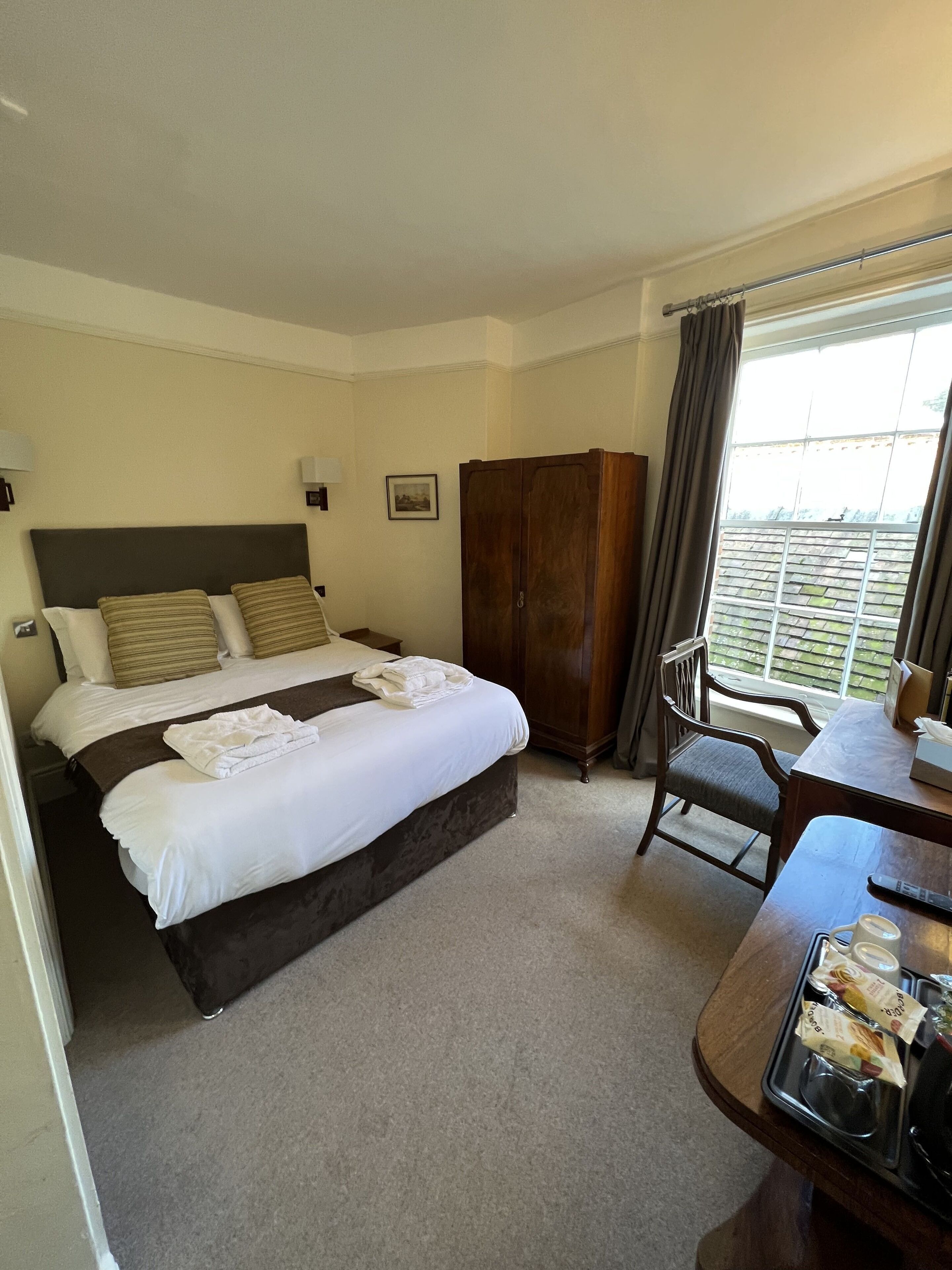 A hotel room with a bed, a wooden wardrobe, a dining table with a chair, and a window with curtains.