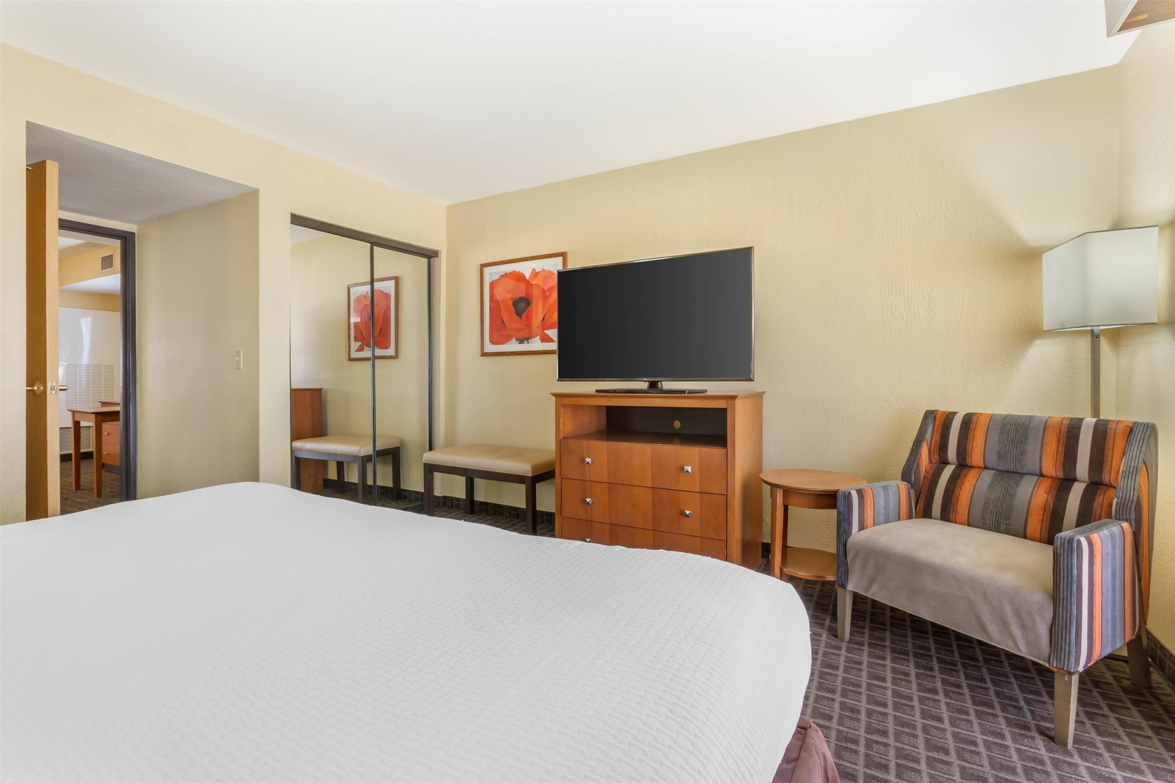 A hotel room with a large bed, a television, a wooden dresser, a striped armchair, and a small table.
