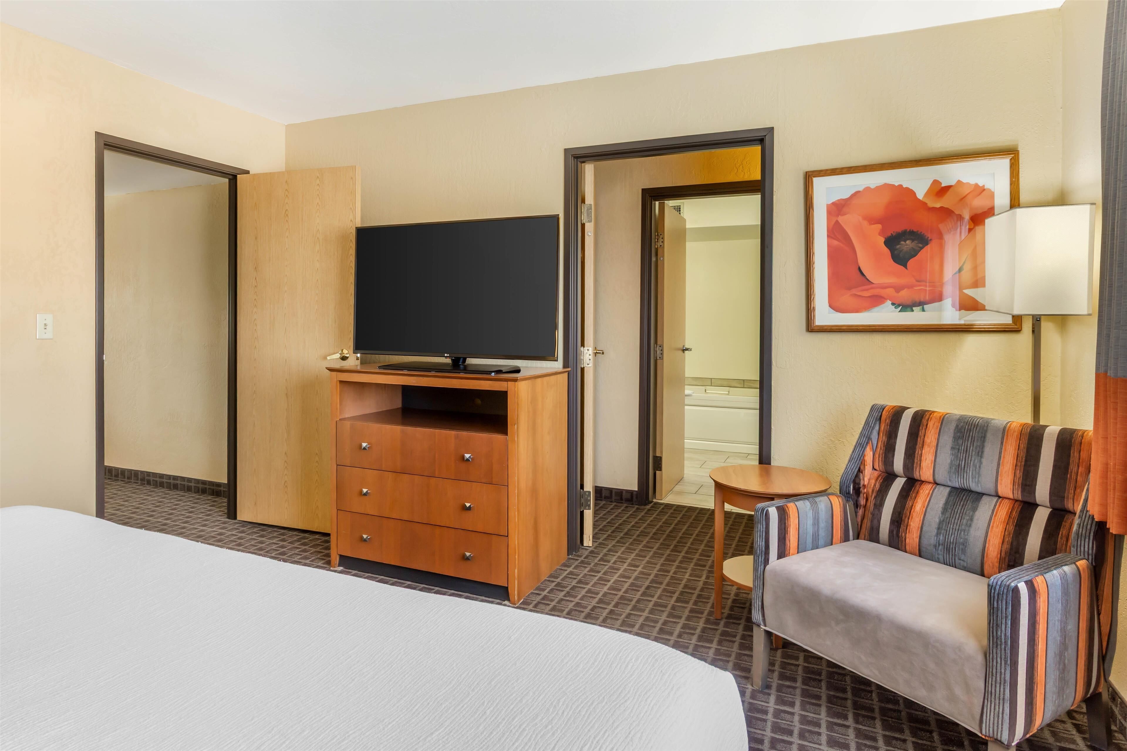 A hotel room with a bed, a wooden dresser with a television, a striped armchair, a small table, and a framed picture on the wall.