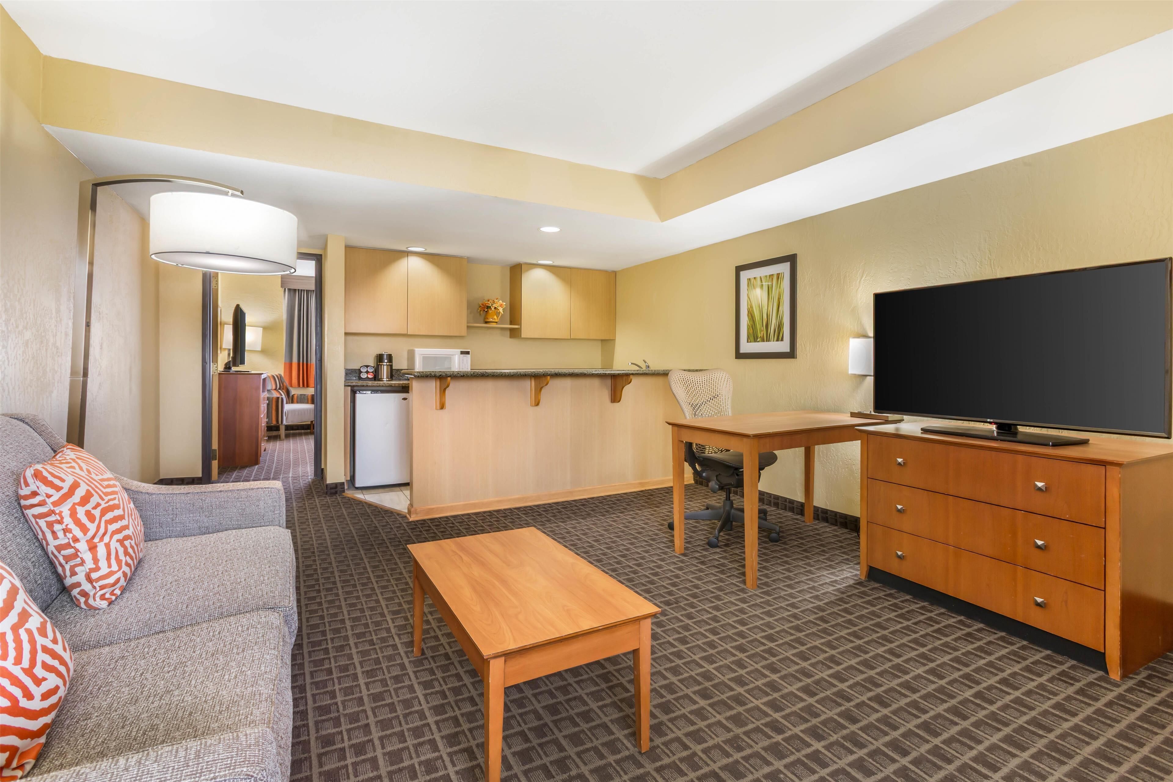 Habitación de hotel con sofá, escritorio de madera, televisor, área de cocina y alfombra estampada.