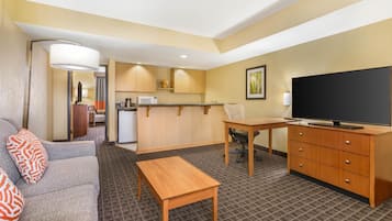 Habitación de hotel con sofá, escritorio de madera, televisor, área de cocina y alfombra estampada.